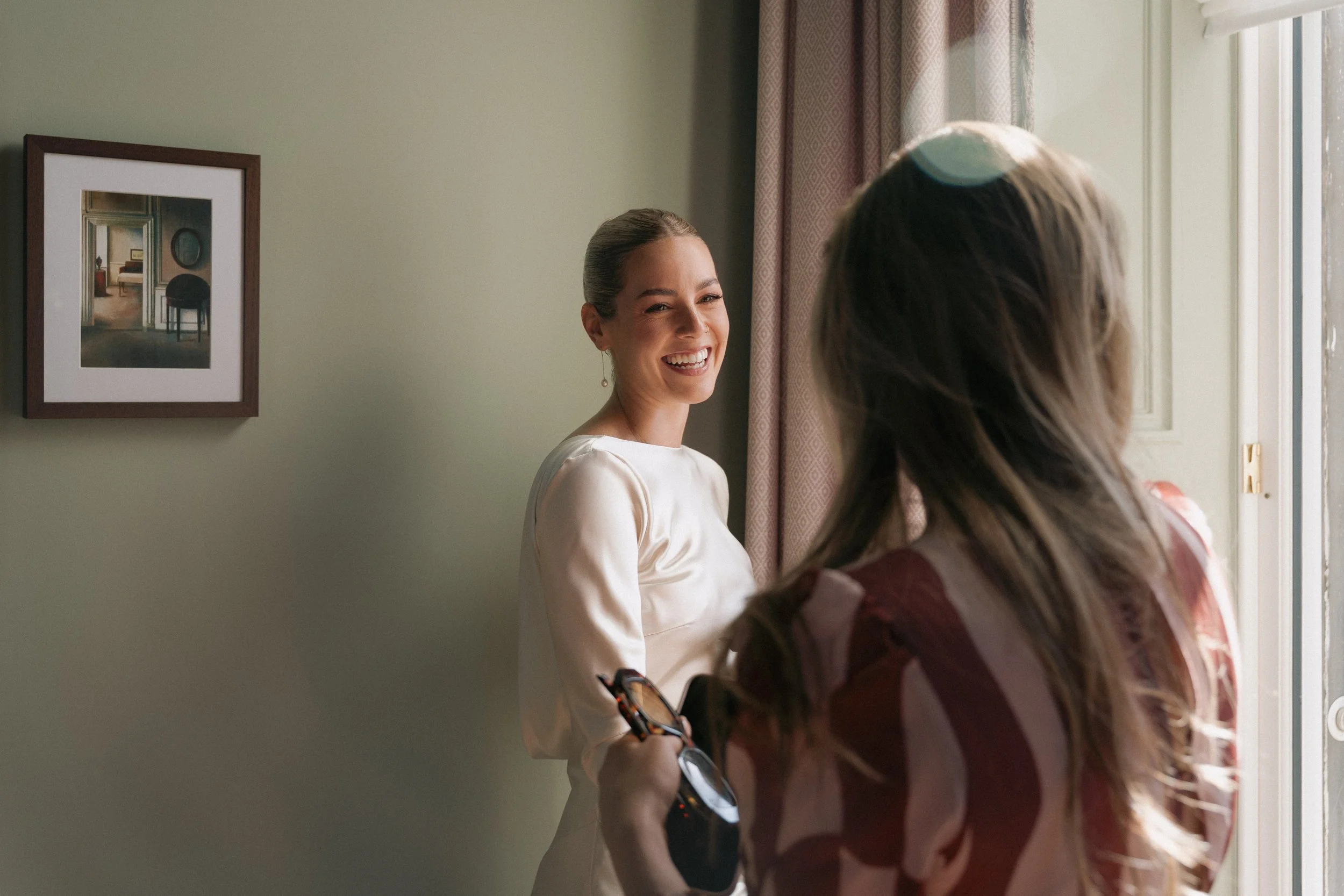 Two women, one smiling, talk near a window with curtains in a home interior. - captured by an Edinburgh wedding photographer