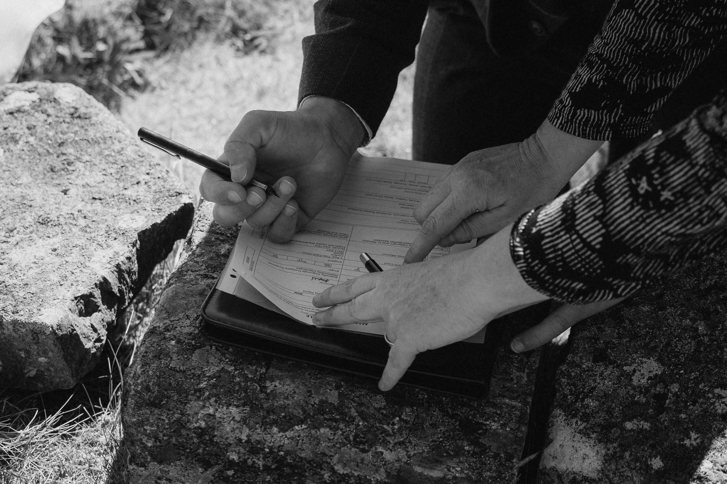 Two people are filling out a form on a clipboard outdoors, with one person pointing at the paper and both holding pens, sitting on rocks. - captured by an Edinburgh wedding photographer