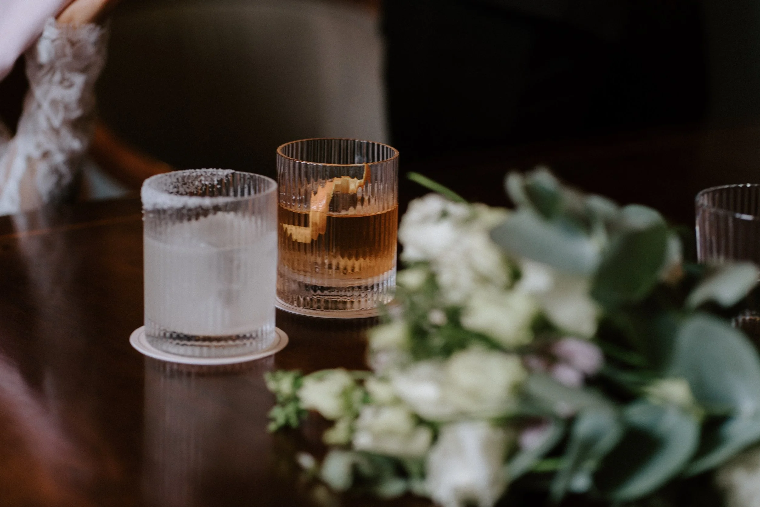 A glass of water with a salt rim, a glass of whiskey with a lemon peel, and a bouquet of white flowers on a wooden table. - captured by an Edinburgh wedding photographer
