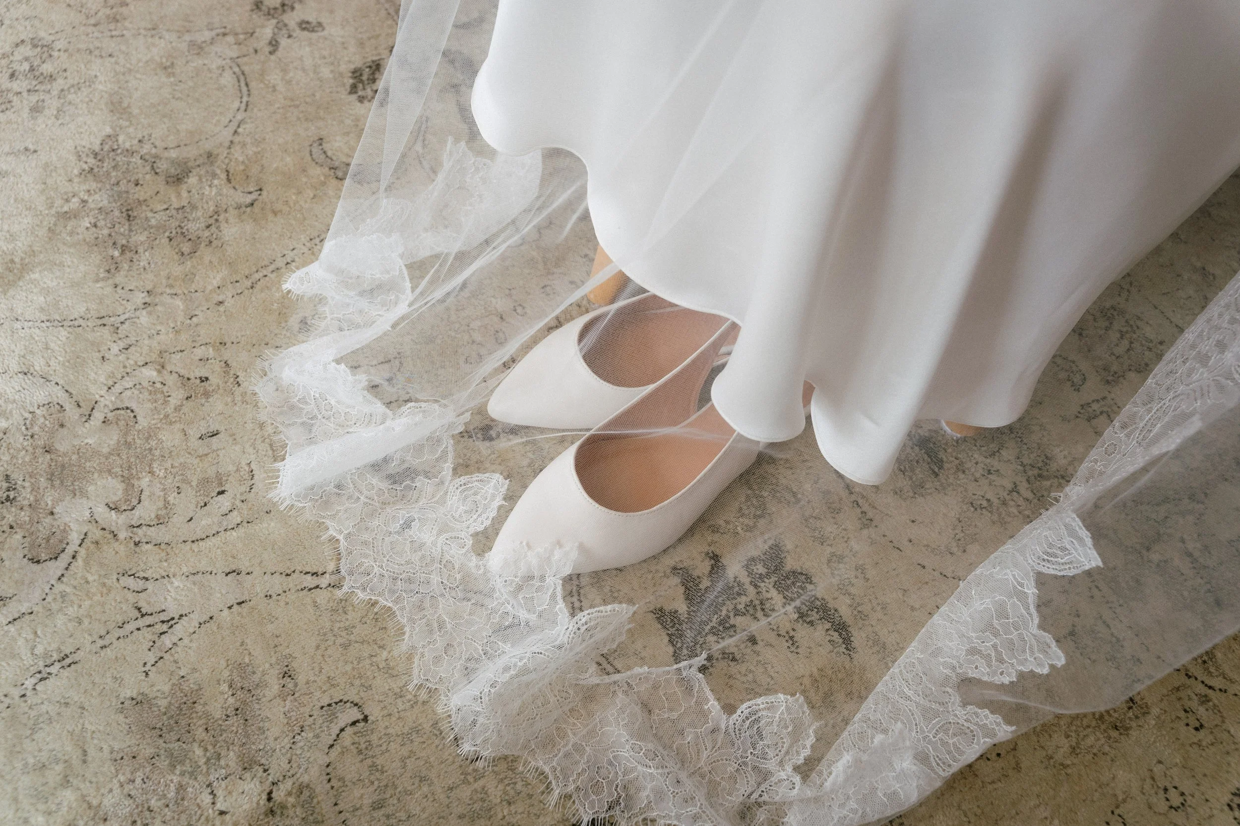 White high-heeled shoes under a white wedding dress with lace hem on a textured carpeted floor. - captured by an Edinburgh wedding photographer