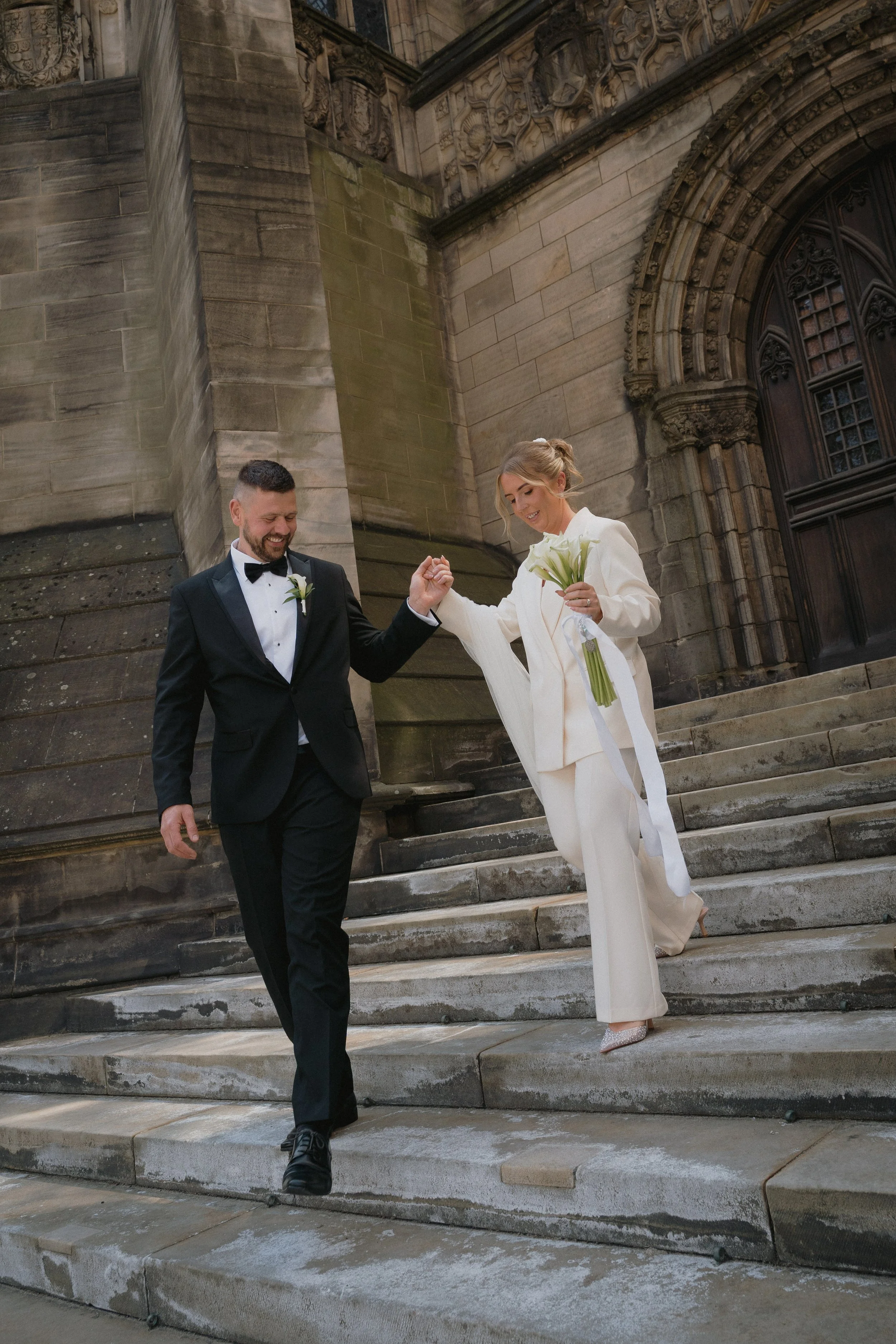 A bride and groom walking down stone steps outside a historic building, smiling and holding hands. - captured by an Edinburgh wedding photographer