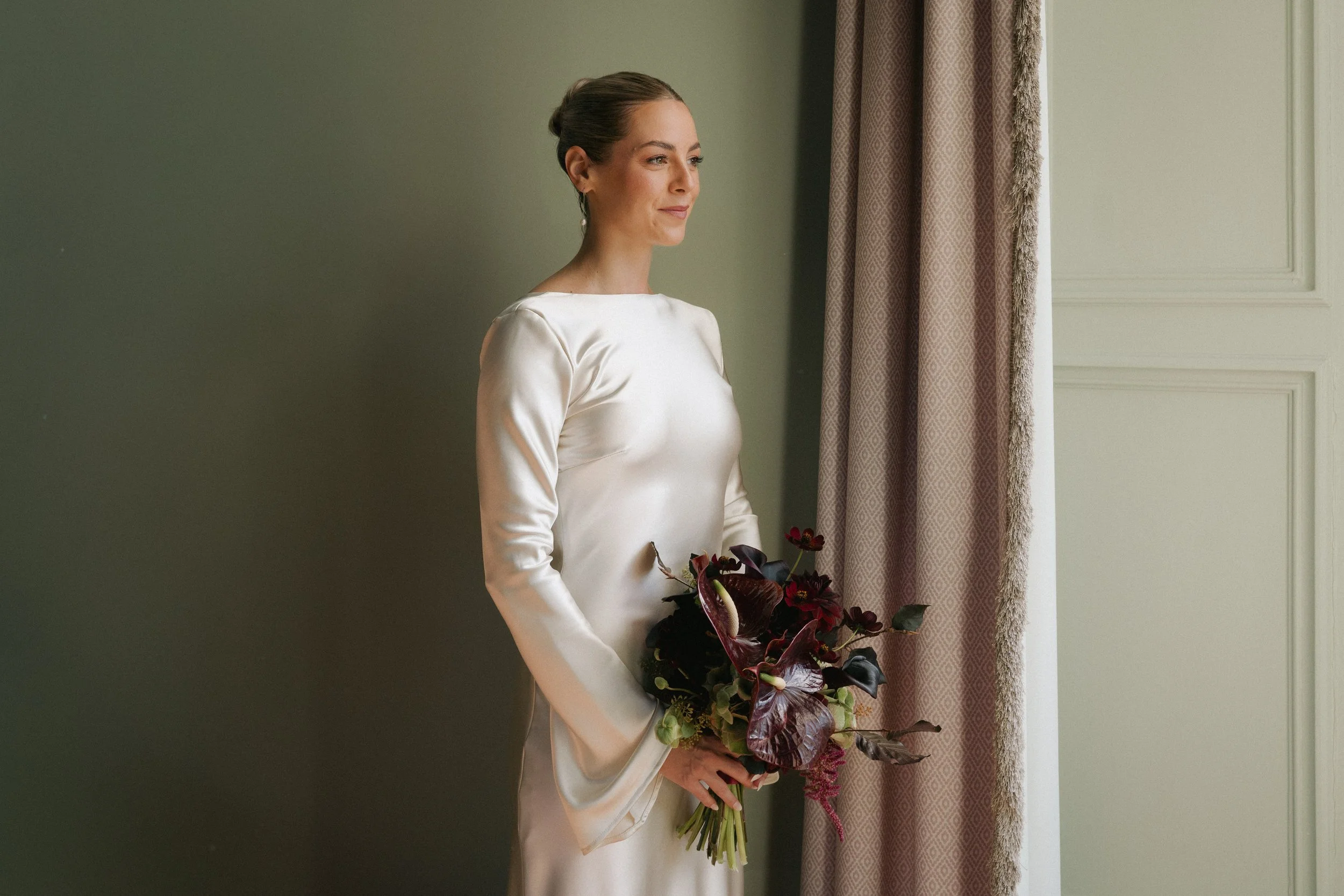 A woman in a white satin dress holding a dark red flower bouquet, standing indoors near a window with gray and pink curtains. - captured by an Edinburgh wedding photographer