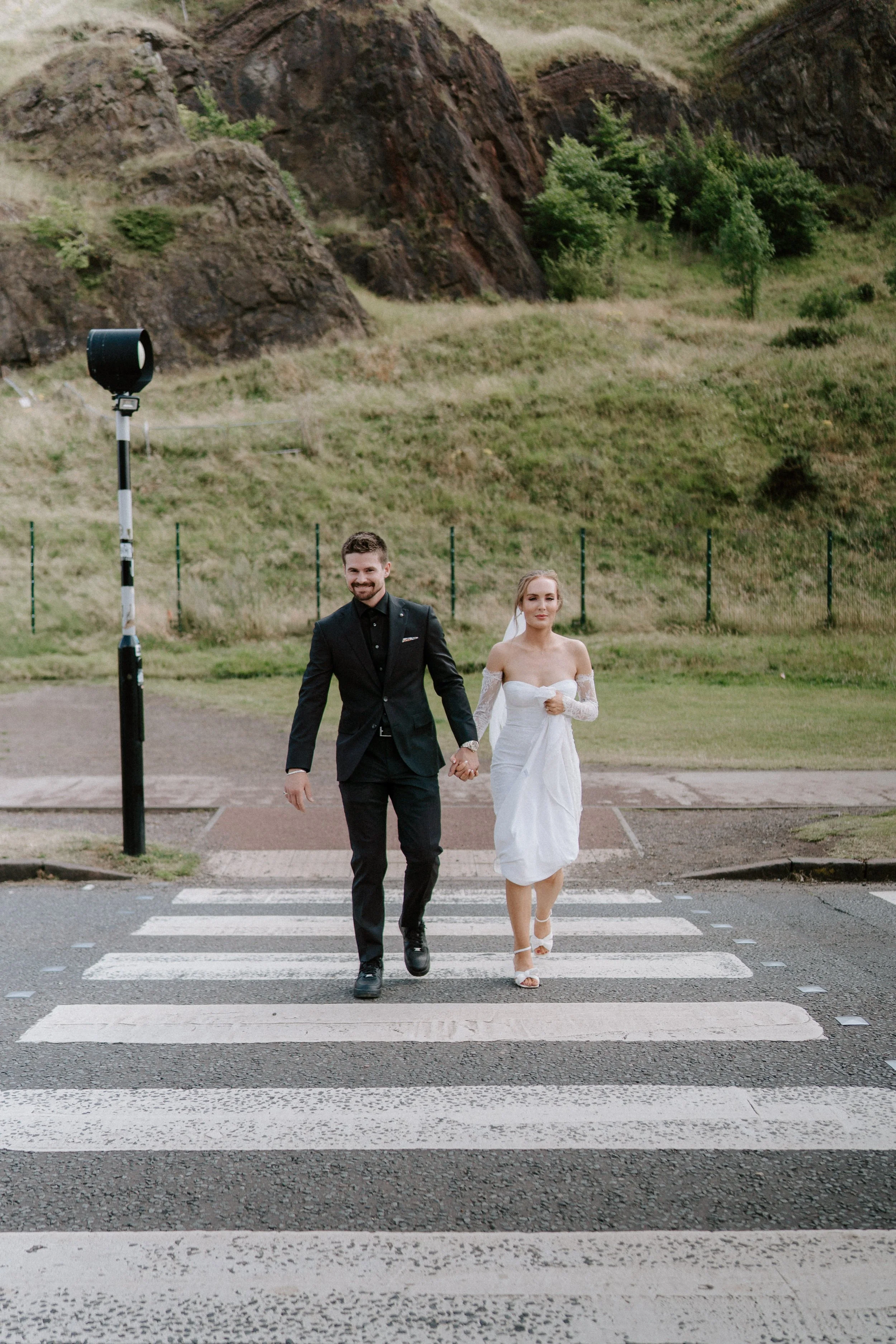 A newlywed couple walking hand-in-hand across a pedestrian crosswalk outdoors, with a rocky hillside and green trees in the background. - captured by an Edinburgh wedding photographer