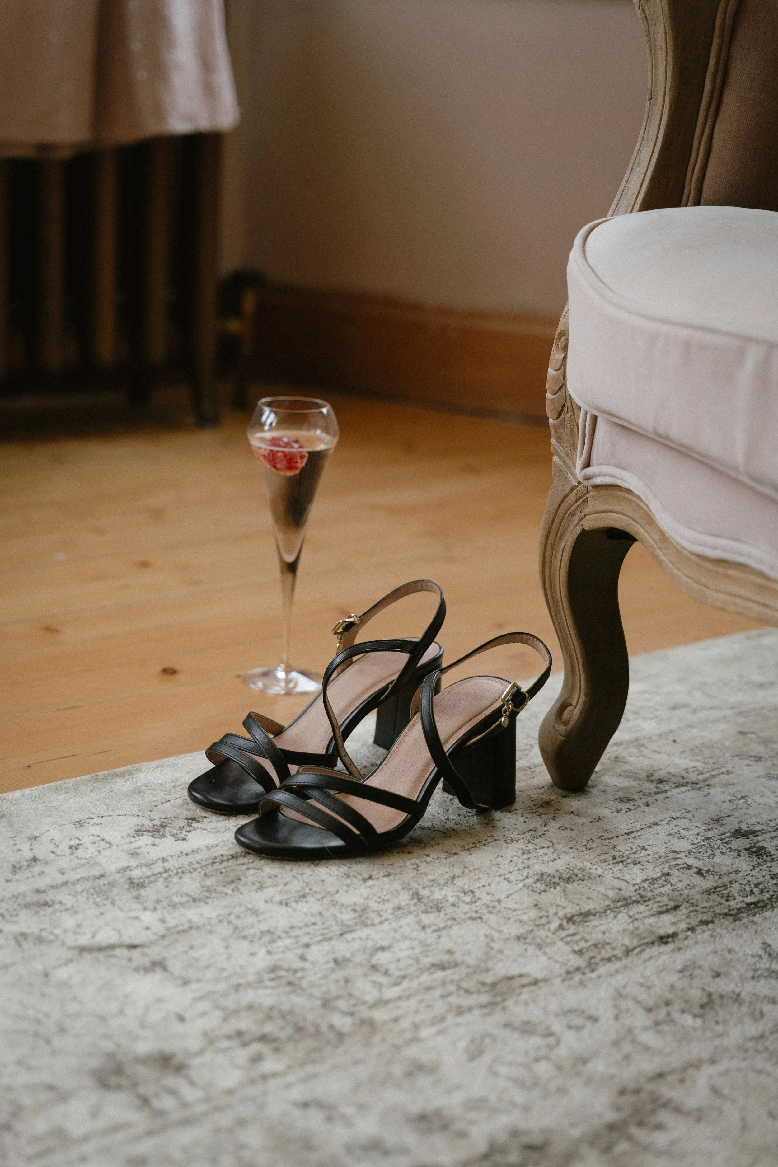 A pair of black high-heeled sandals and a glass of red wine with berries on a patterned rug next to an elegant upholstered armchair. - captured by an Edinburgh wedding photographer