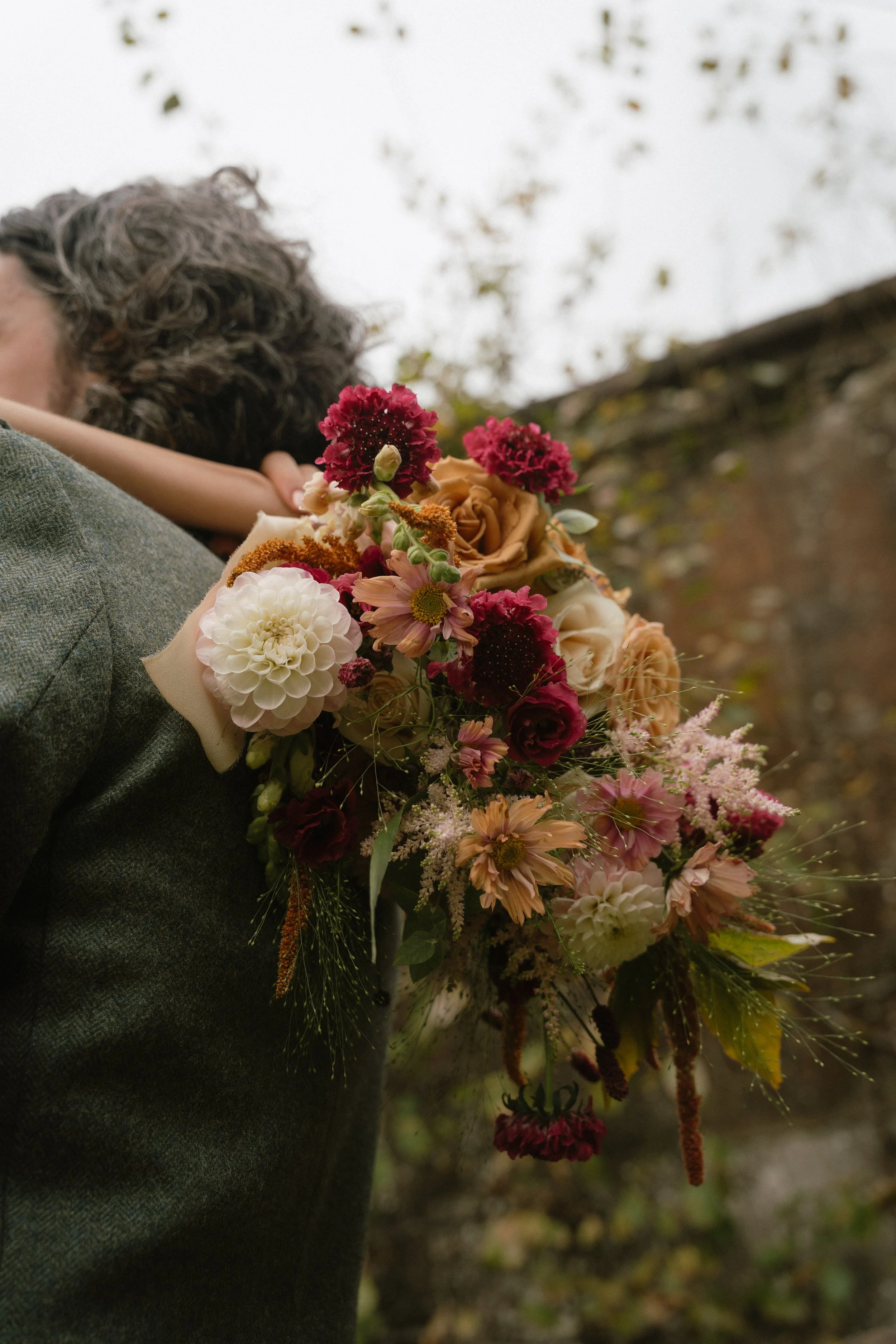 A person in a grey suit jacket holding a colorful bouquet of flowers with pink, white, and peach blossoms, outdoors against a blurred background of trees and cloudy sky. - captured by an Edinburgh wedding photographer