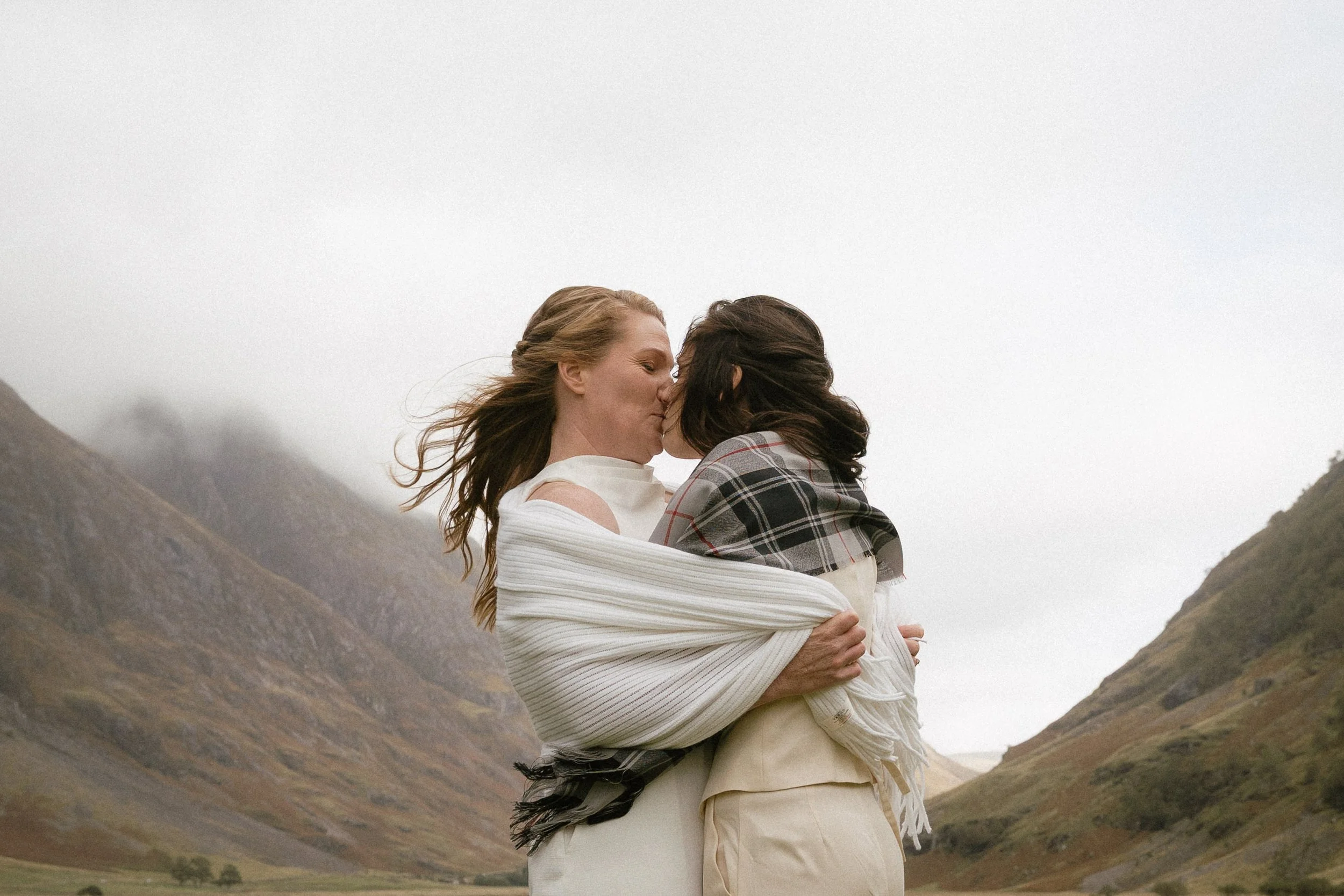 Two women embrace and kiss outdoors in a mountainous landscape, with overcast sky and rolling hills in the background. - captured by an Edinburgh wedding photographer