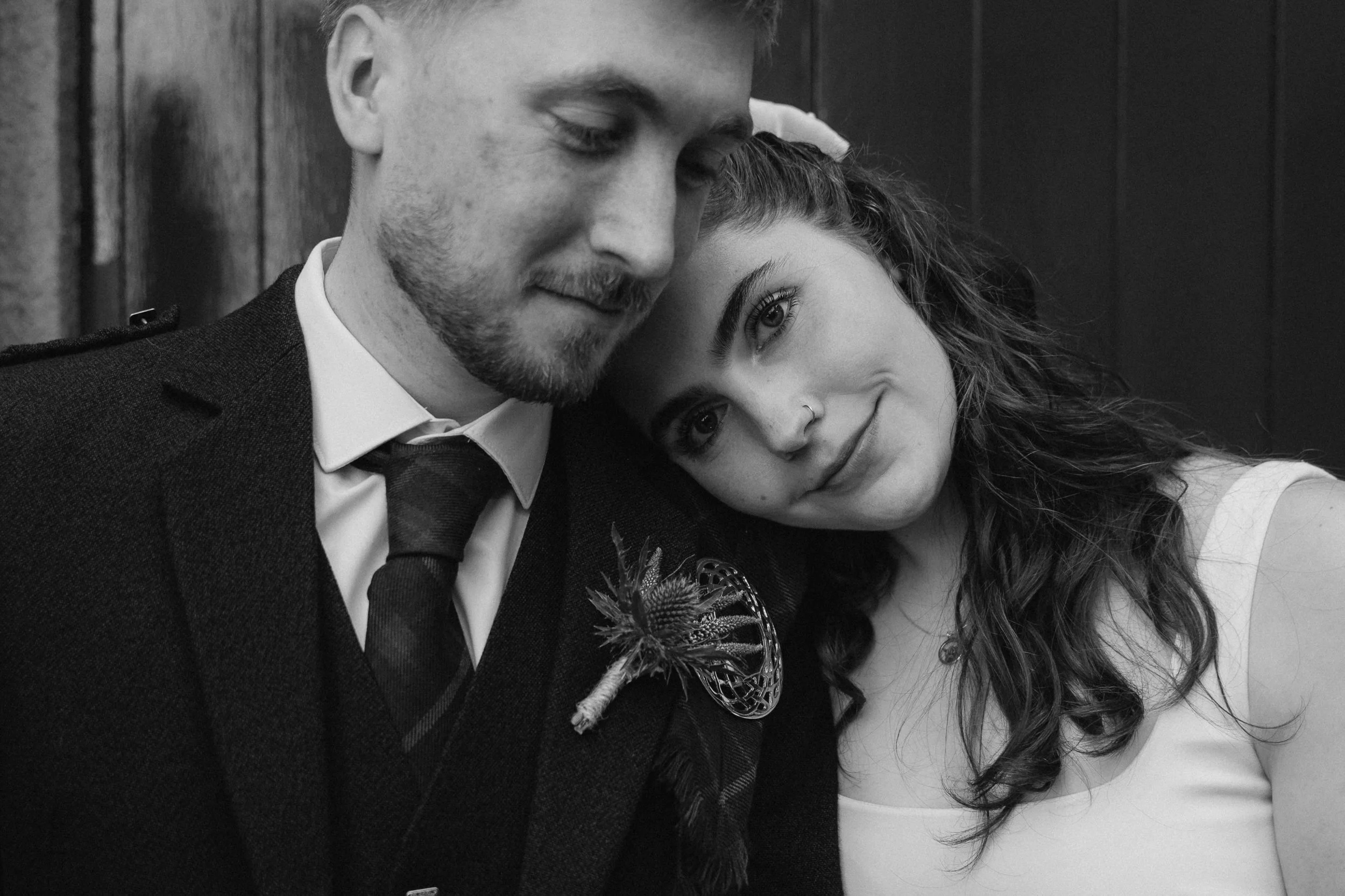 A man and woman with their heads leaning together in a close, affectionate pose. The man is in a suit with a tie and a decorative boutonniere, and the woman has long, curly hair, a nose piercing, and is wearing a sleeveless top. They are smiling soft