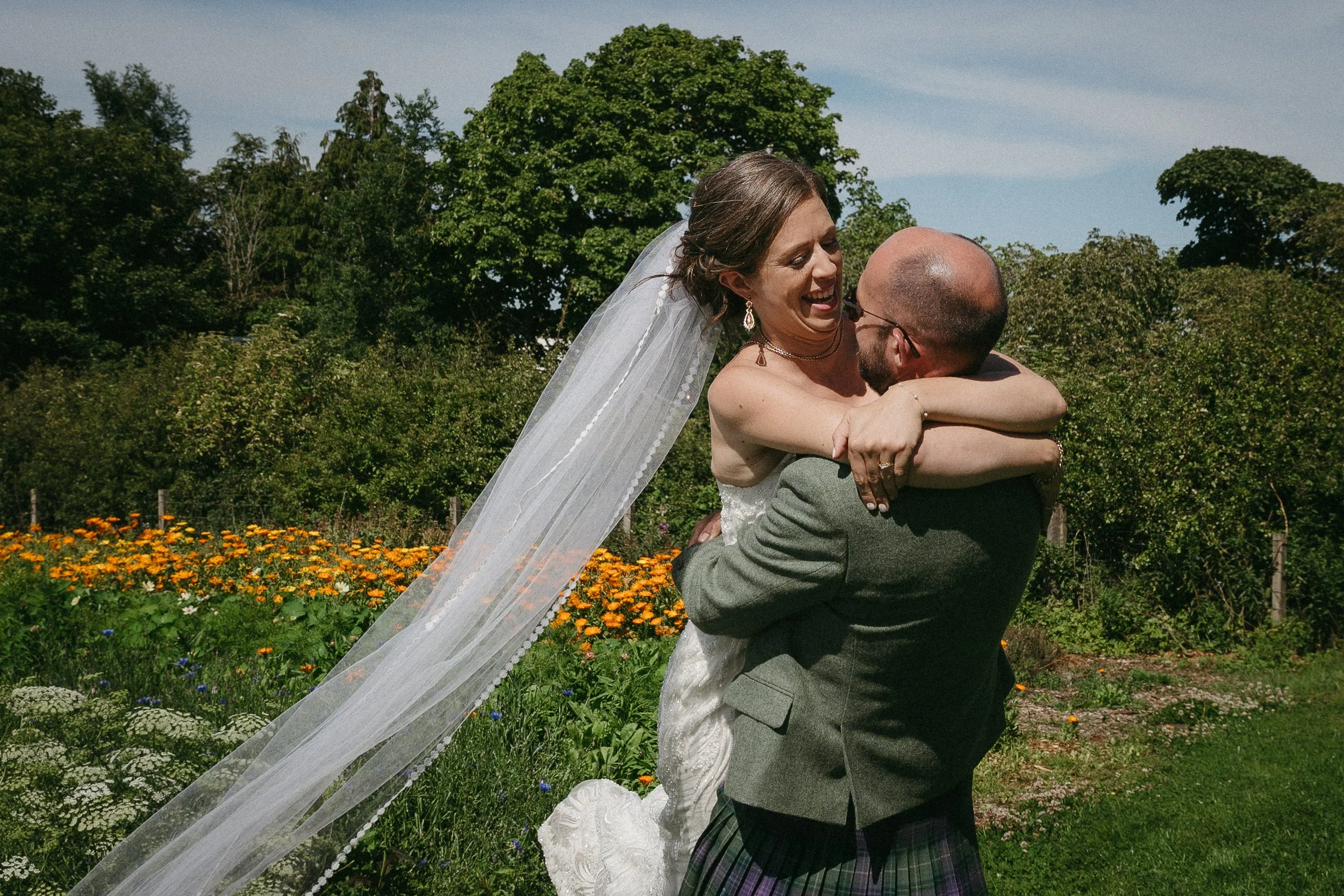 A bride in a white wedding dress and veil is being lifted by a man in a gray suit and plaid kilt, holding her in a garden with colorful flowers and green trees under a blue sky. - captured by an Edinburgh wedding photographer