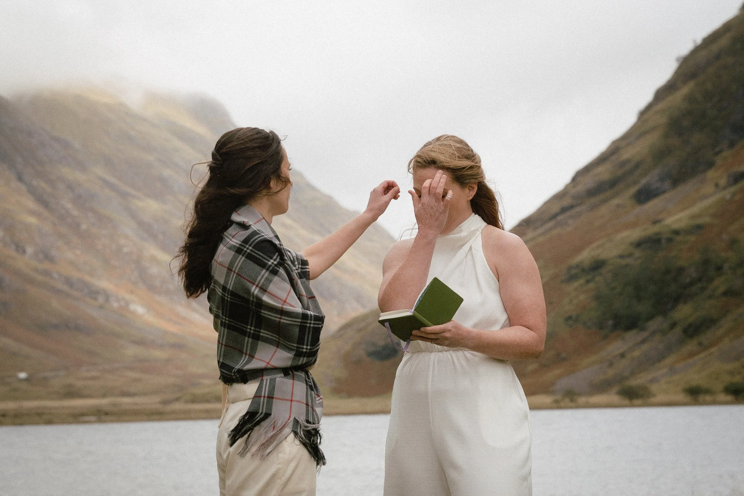 Two women standing beside a lake with mountain scenery in the background; one woman is applying makeup or adjusting her glasses while the other holds a small book or journal.