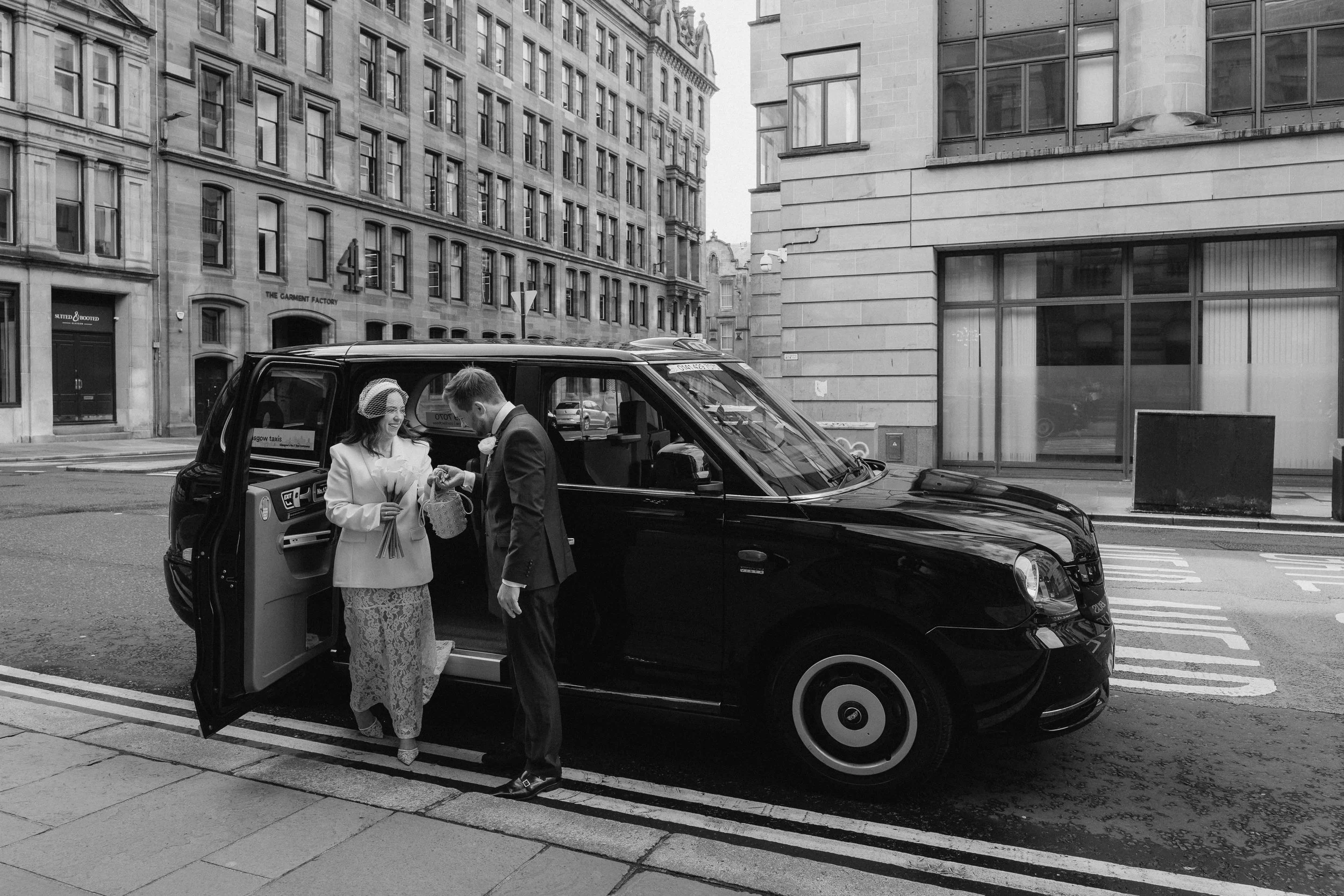 A woman and a man stand outside a black taxi on a city street. The woman is dressed in a lace skirt and a coat, holding a bouquet and a basket, and the man is in a suit with a boutonniere, possibly a wedding scene. - captured by an Edinburgh wedding 