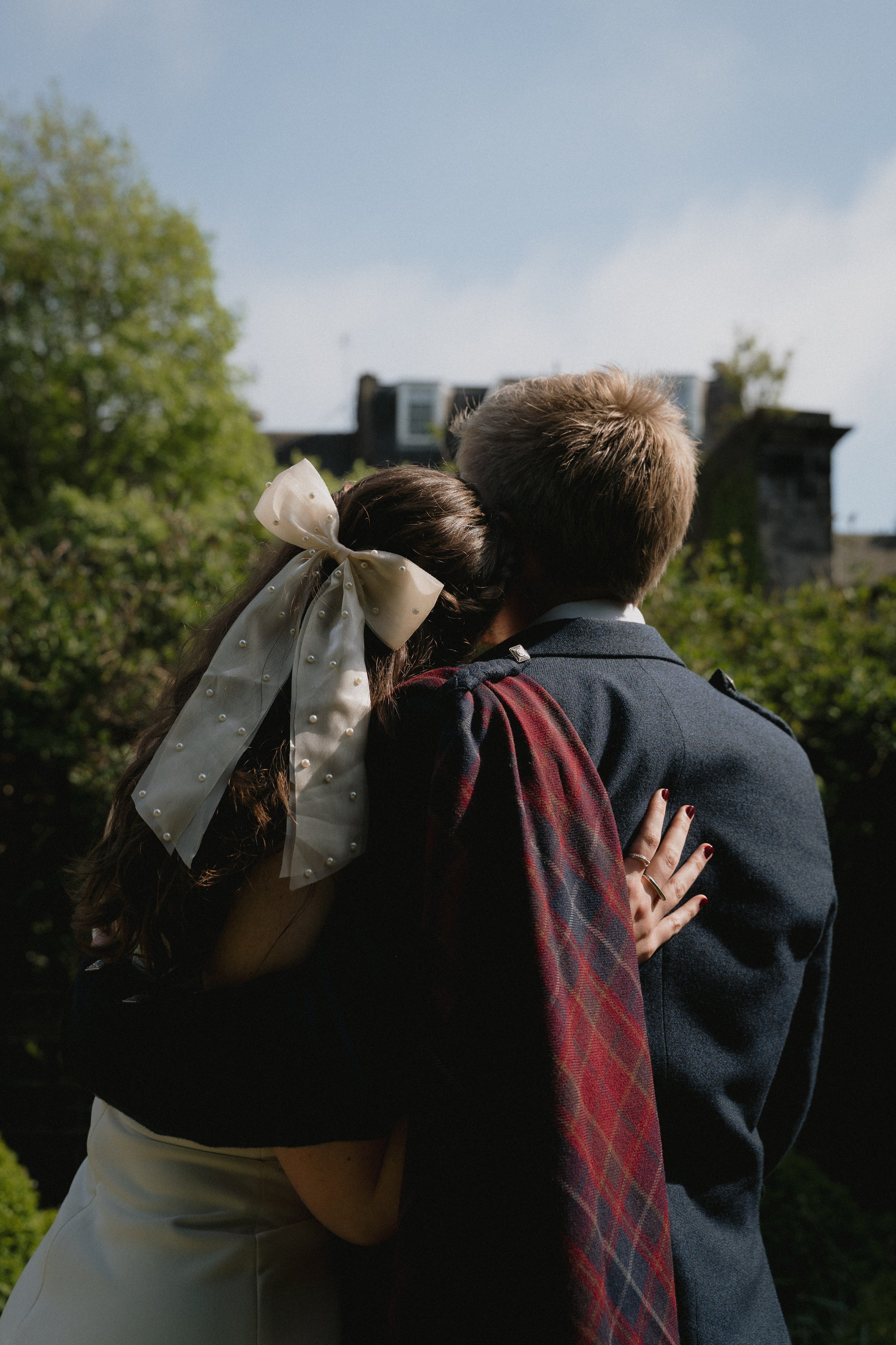 A couple embracing outdoors, viewed from behind, with the woman resting her head on the man's shoulder. - captured by an Edinburgh wedding photographer