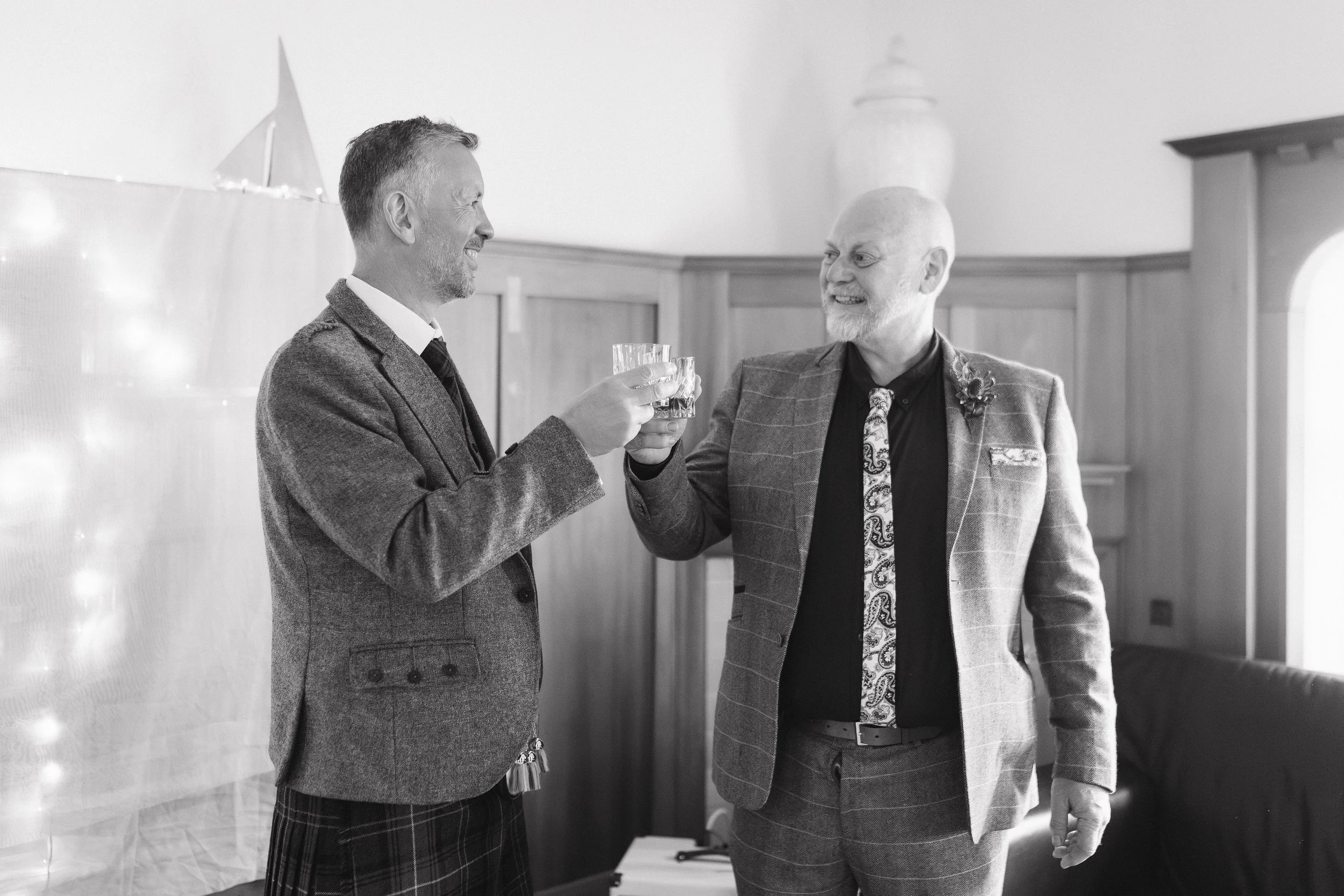 Two men in suits toasting with glasses in a room with wood paneling. - captured by an Edinburgh wedding photographer