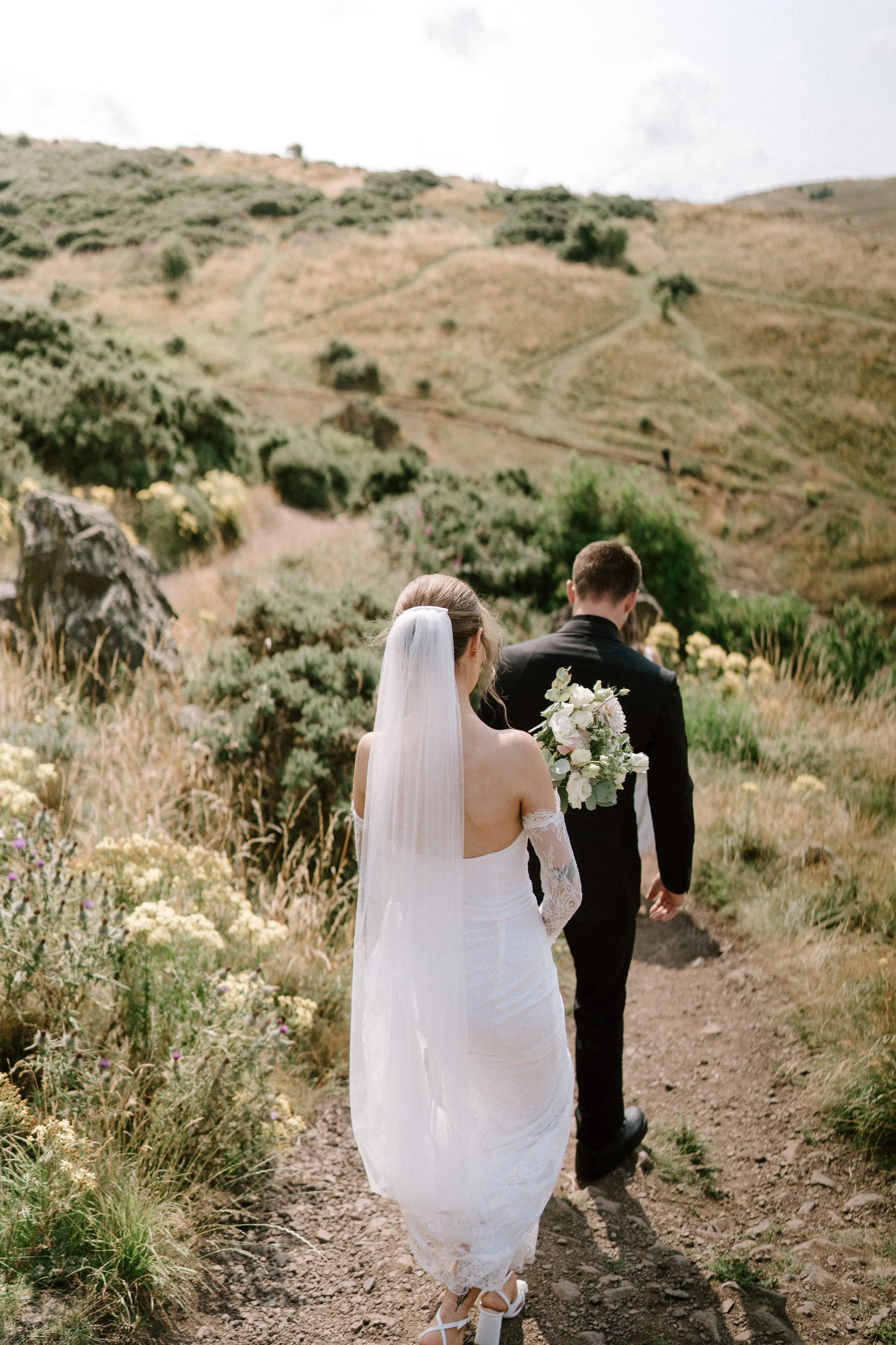 A bride and groom walking on a dirt path through a grassy, hilly landscape with shrubs and flowers, seen from behind. - captured by an Edinburgh wedding photographer