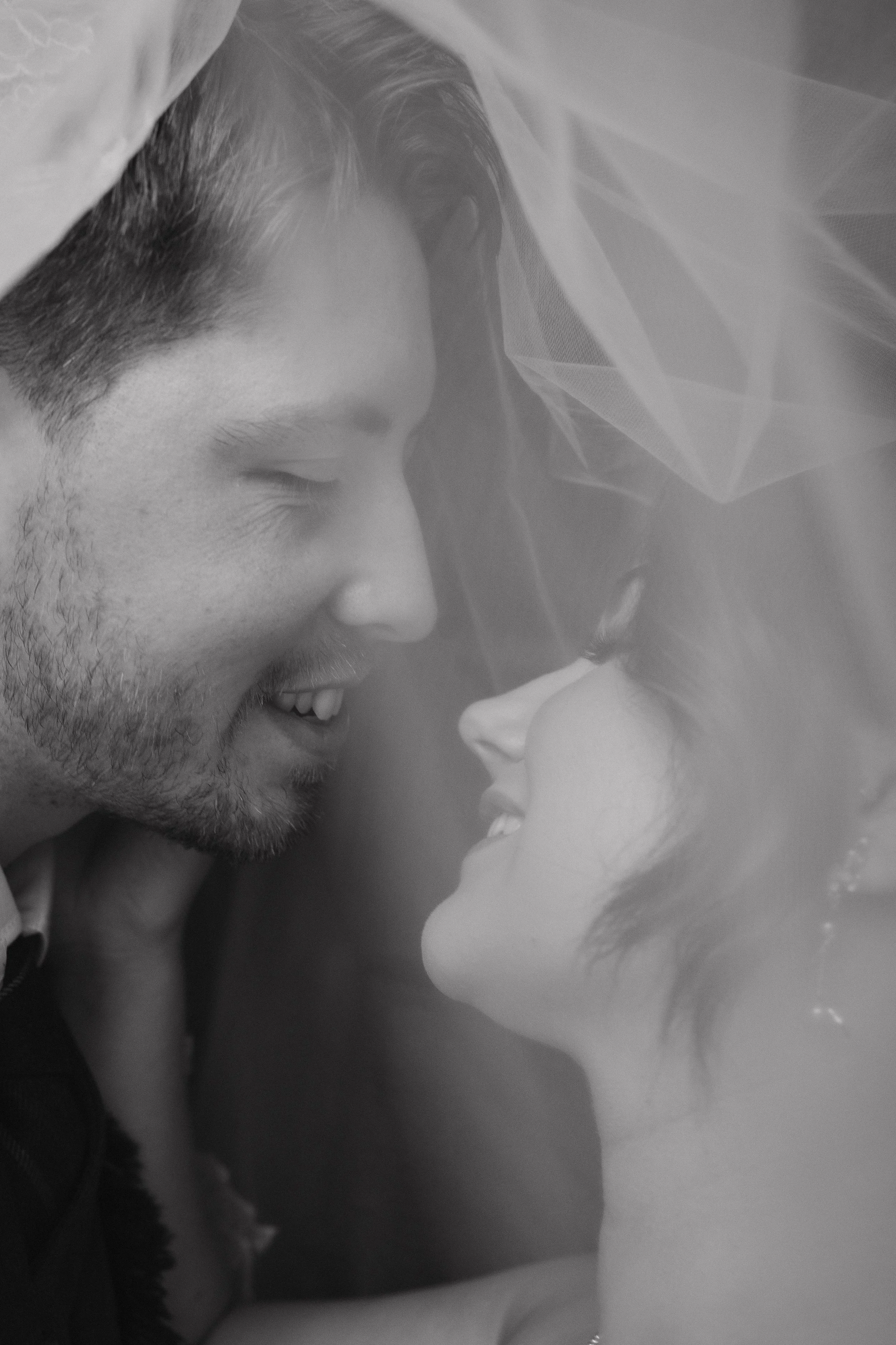 Black and white photo of a smiling man and woman with their faces close together under a veil, sharing an intimate moment. - captured by an Edinburgh wedding photographer