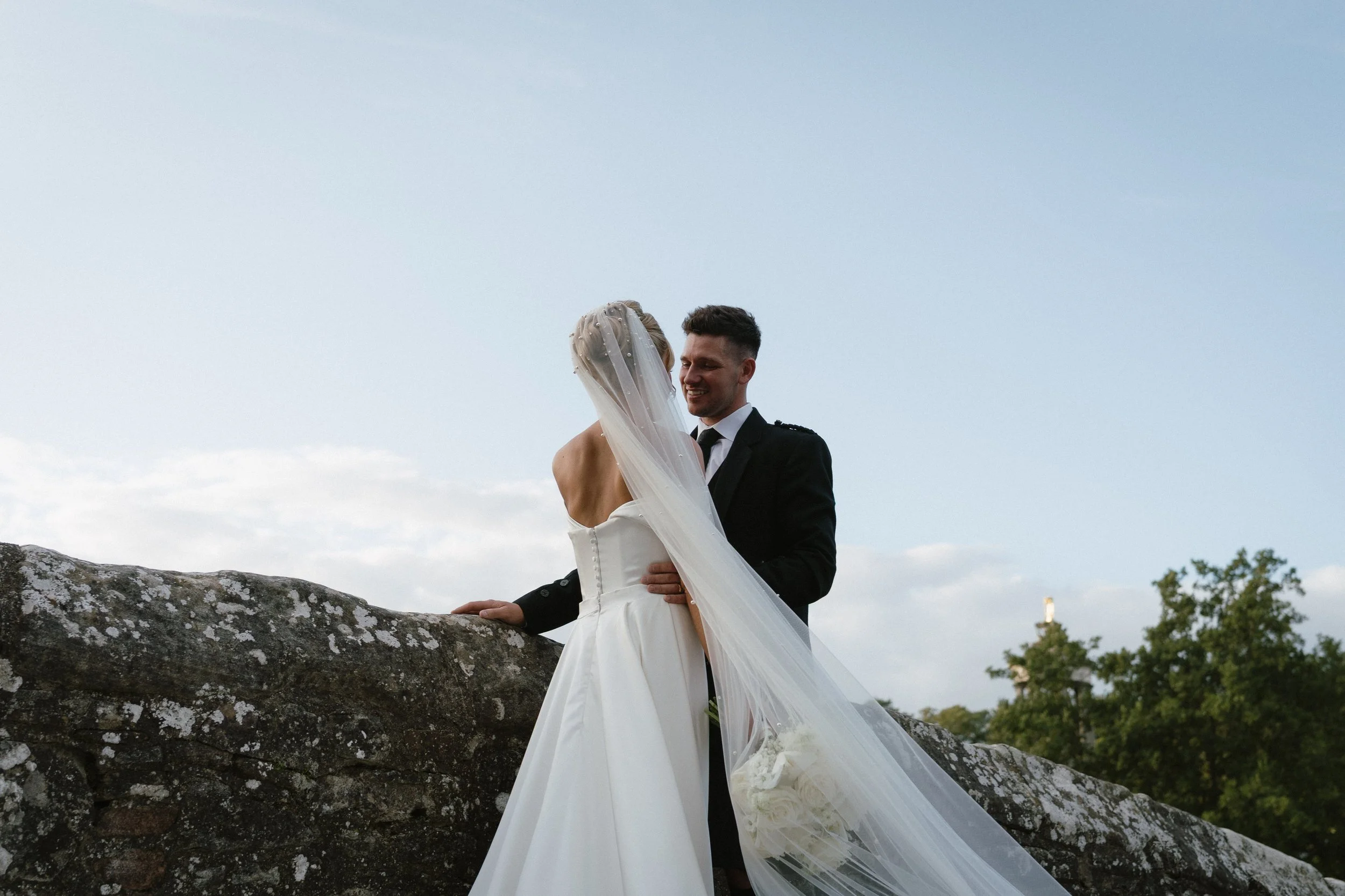 A bride and groom share a moment outdoors on their wedding day, leaning against a stone wall with trees and a cloudy sky in the background. - captured by an Edinburgh wedding photographer