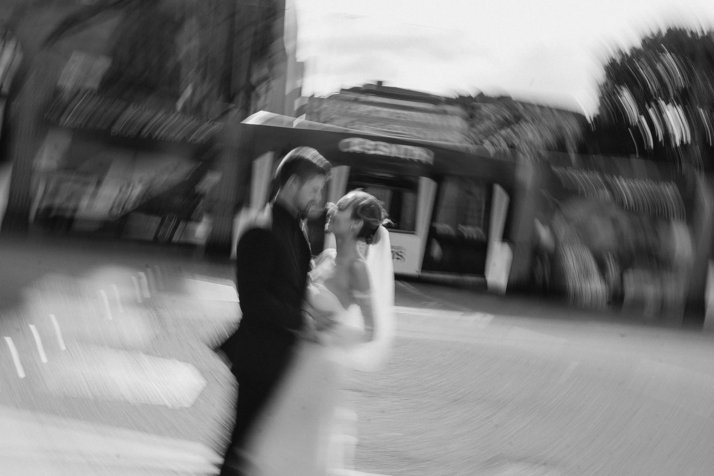 Black and white photo of a bride and groom sharing a kiss on the street, with a bus and buildings in the background. The image appears motion blurred. - captured by an Edinburgh wedding photographer