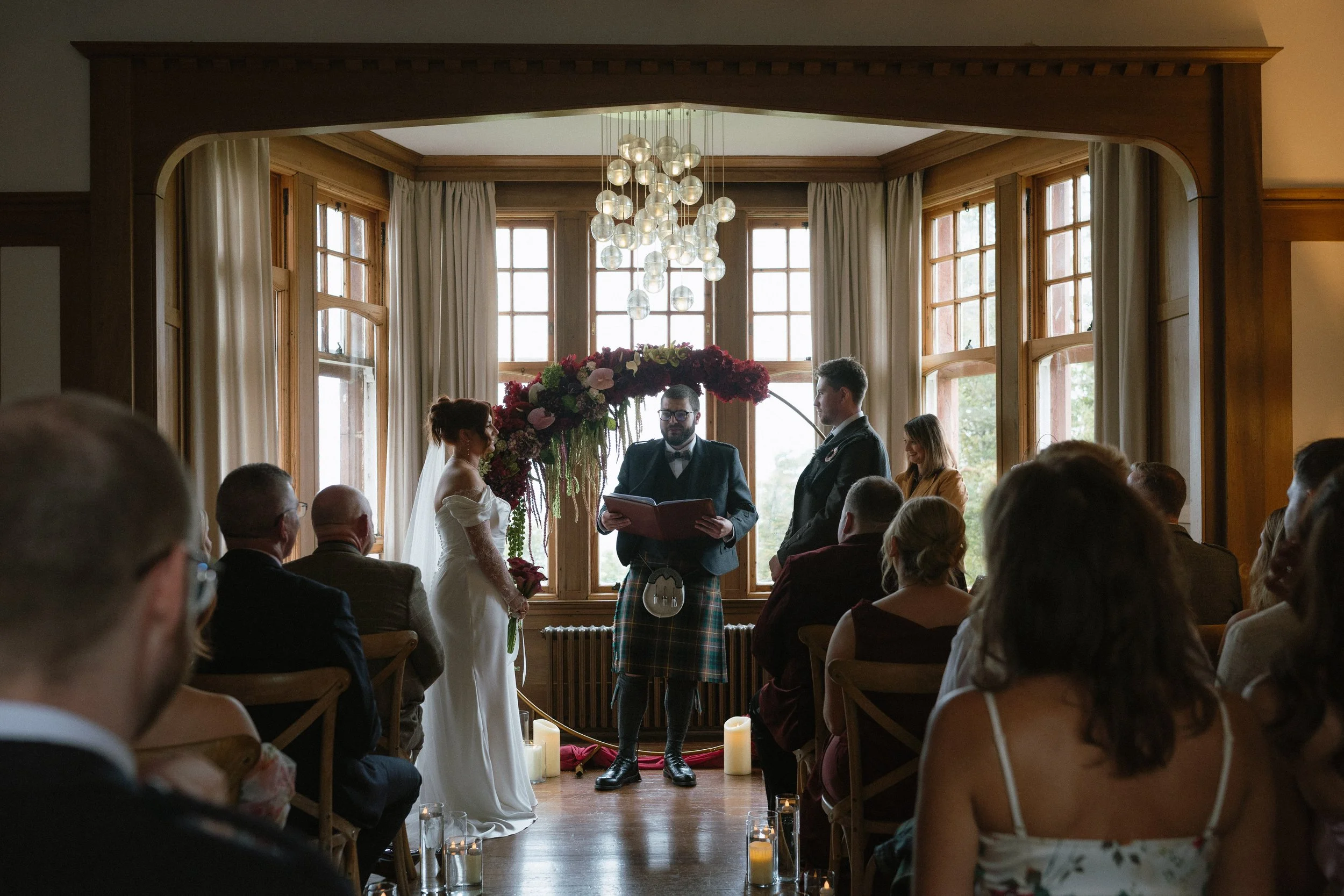A wedding ceremony with a bride and groom standing at the altar, exchanging vows. The officiant reads from a book, and there is floral decor above them. Guests are seated, watching the ceremony in a warmly lit room with large windows and candles on t