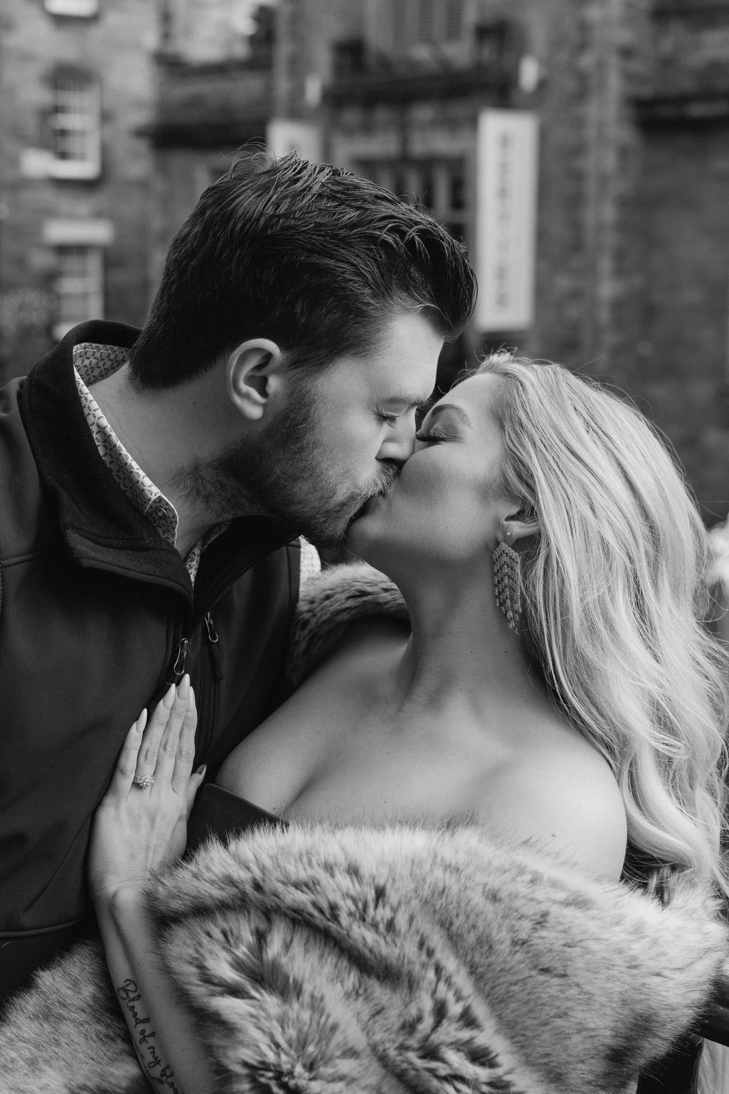 A black and white photograph of a couple sharing a kiss outdoors. The man has dark hair and a beard, and the woman has long blonde hair and wears earrings. They are dressed warmly, with the woman in a fur stole and the man in a jacket. The woman show