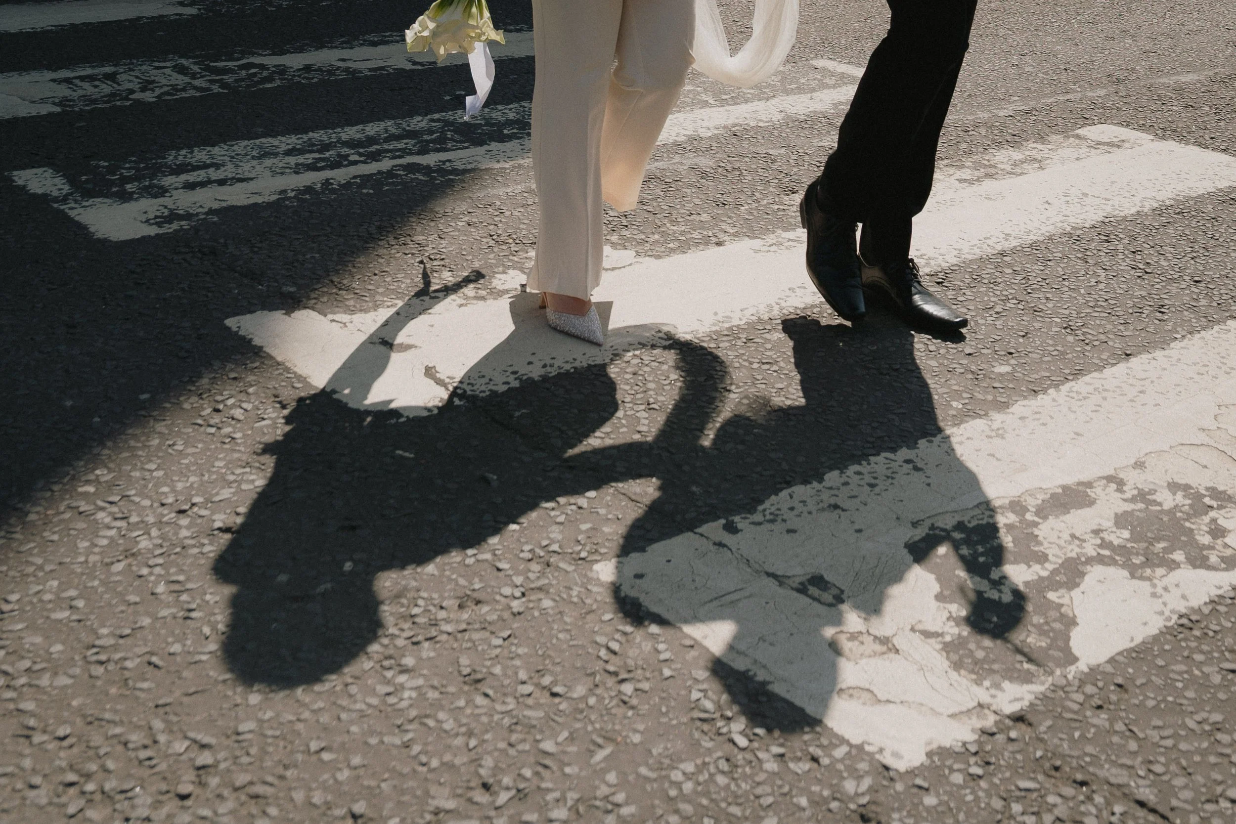 Shadows of a couple holding hands while crossing a street at a pedestrian crosswalk, with one person wearing white shoes and white suit and the other wearing black shoes and black pants. - captured by an Edinburgh wedding photographer