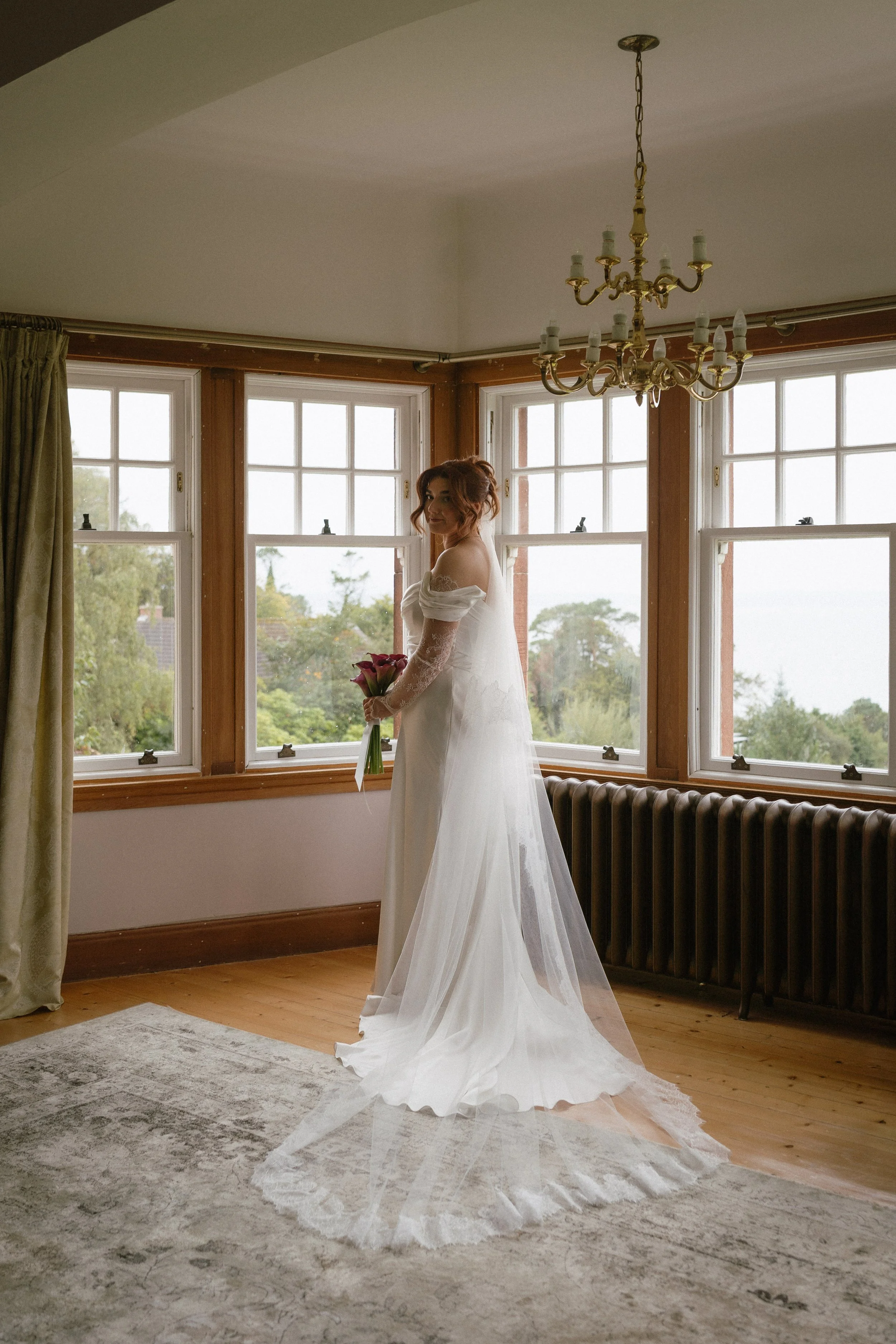 A bride in a white wedding dress holding a bouquet of flowers standing in a room with large windows and a chandelier. - captured by an Edinburgh wedding photographer
