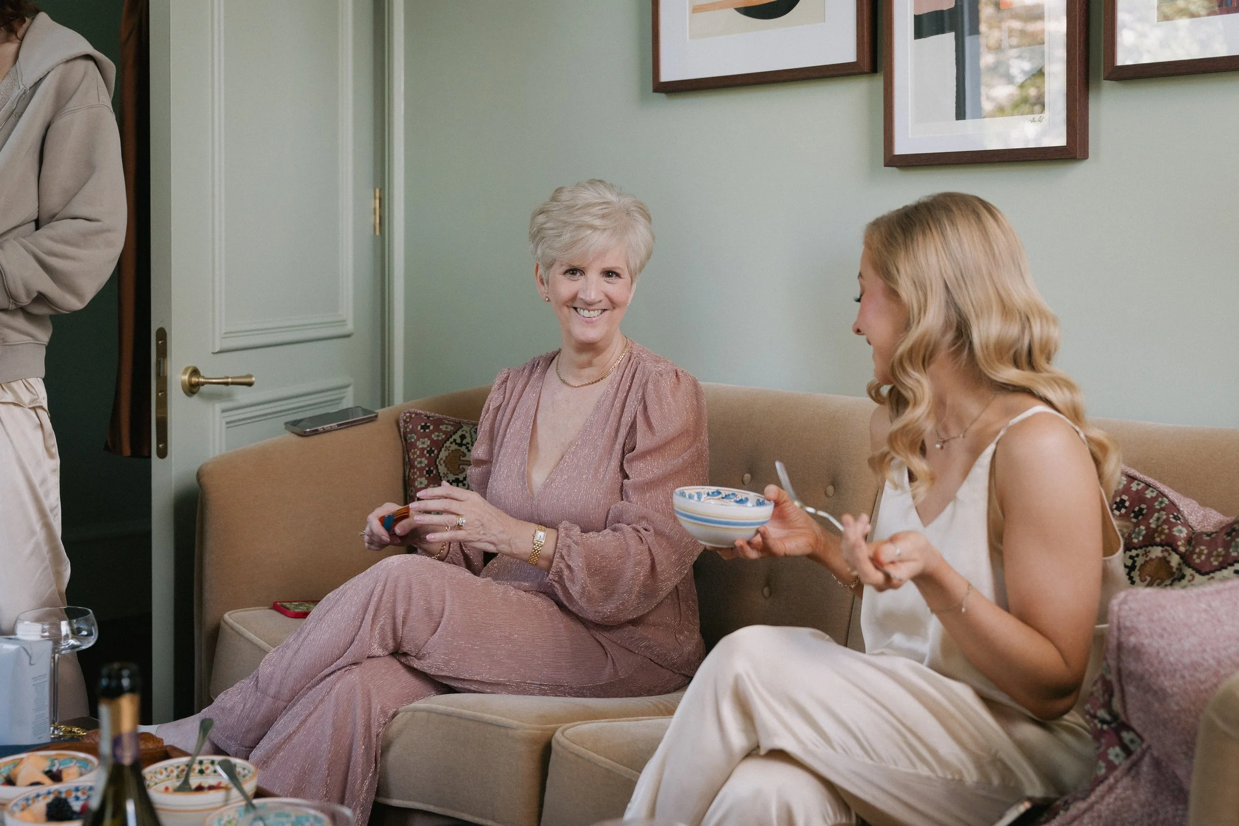 Two women sitting on a beige sofa, smiling and having a conversation during a social gathering in a living room with framed artwork on the wall. - captured by an Edinburgh wedding photographer