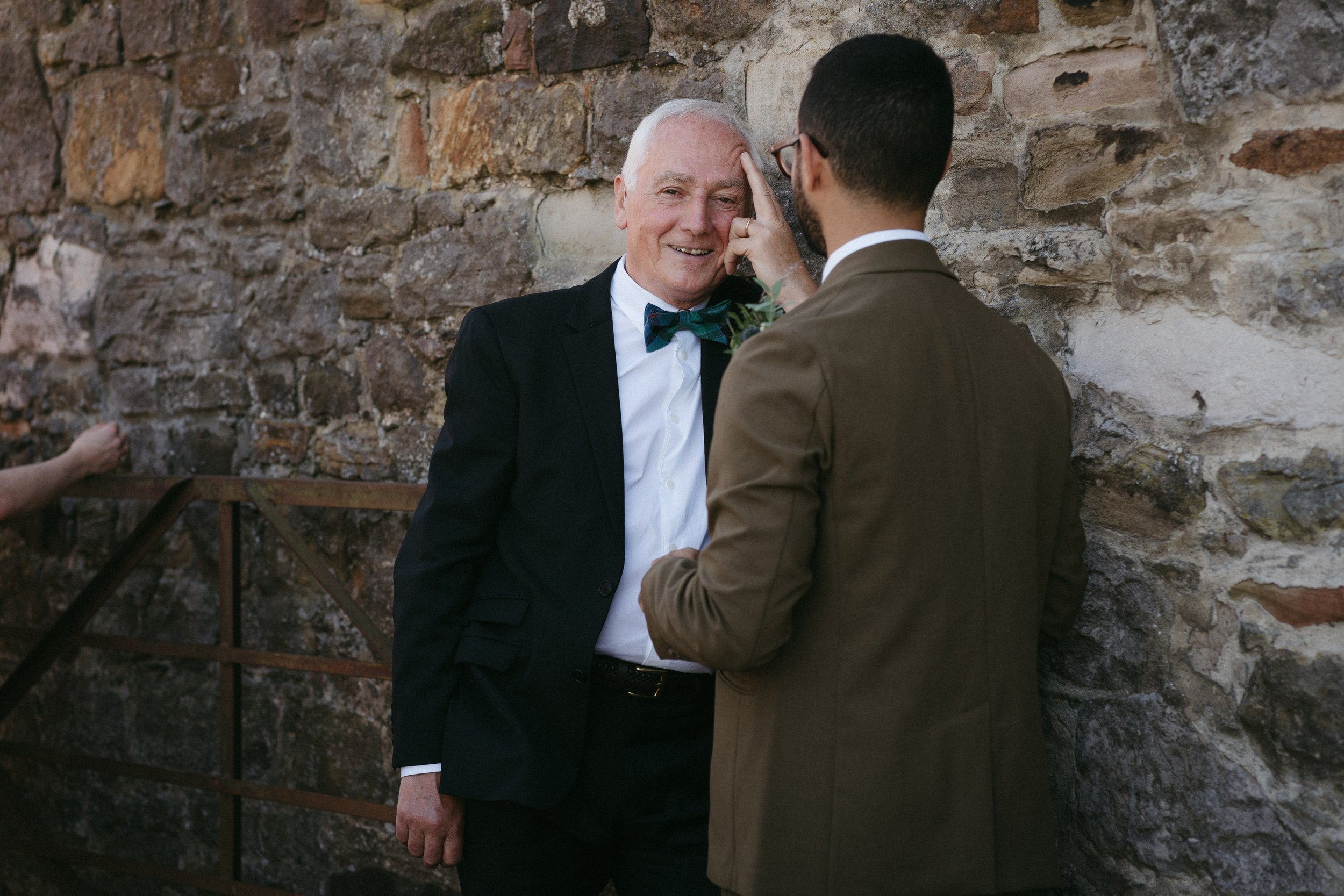 A man in a tuxedo with a bow tie smiling as another man touches his forehead against a stone wall. - captured by an Edinburgh wedding photographer