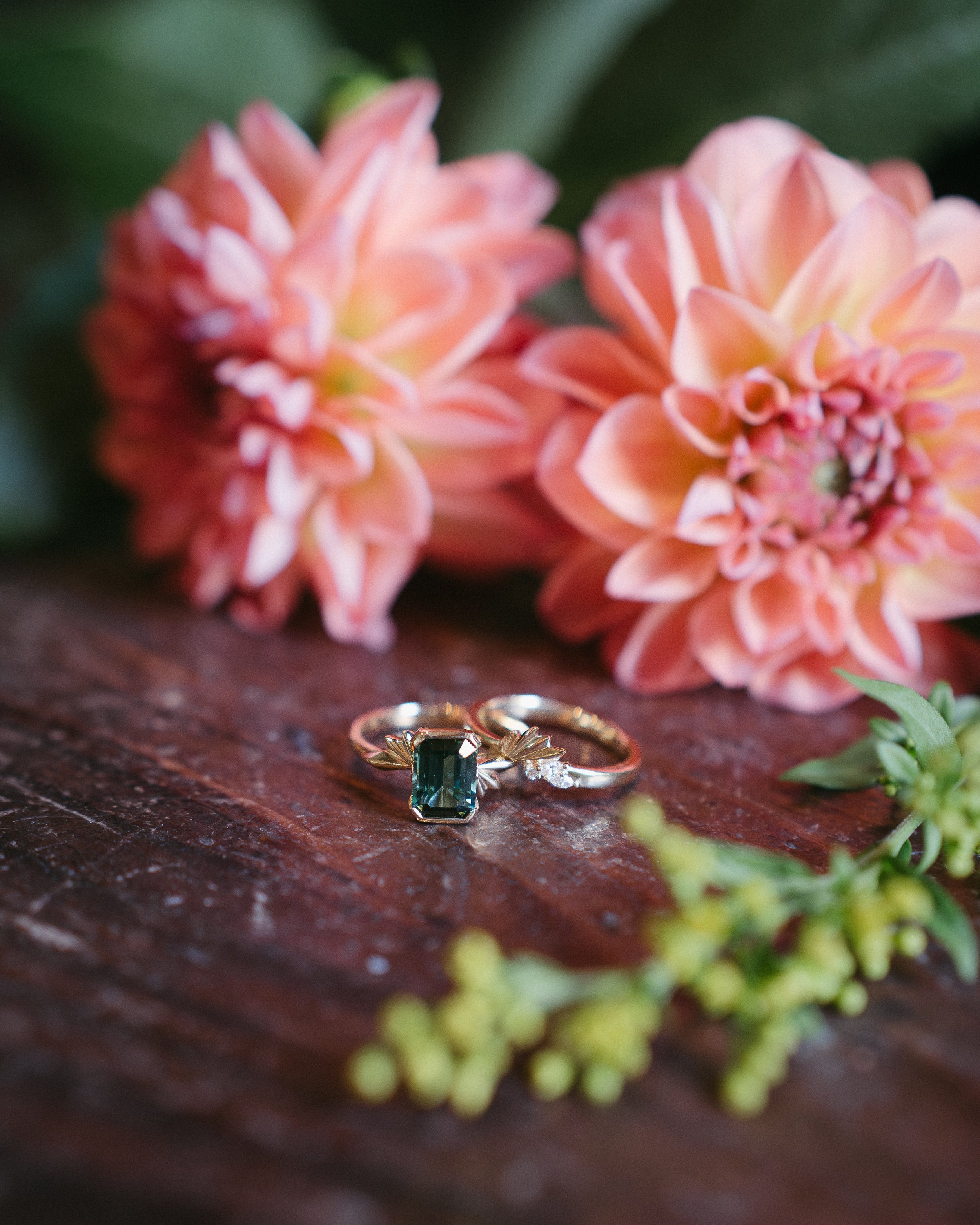 Two rings, one with a rectangular green gemstone and the other with small diamonds, are placed on a wooden surface surrounded by pink flowers and green plants. - captured by an Edinburgh wedding photographer