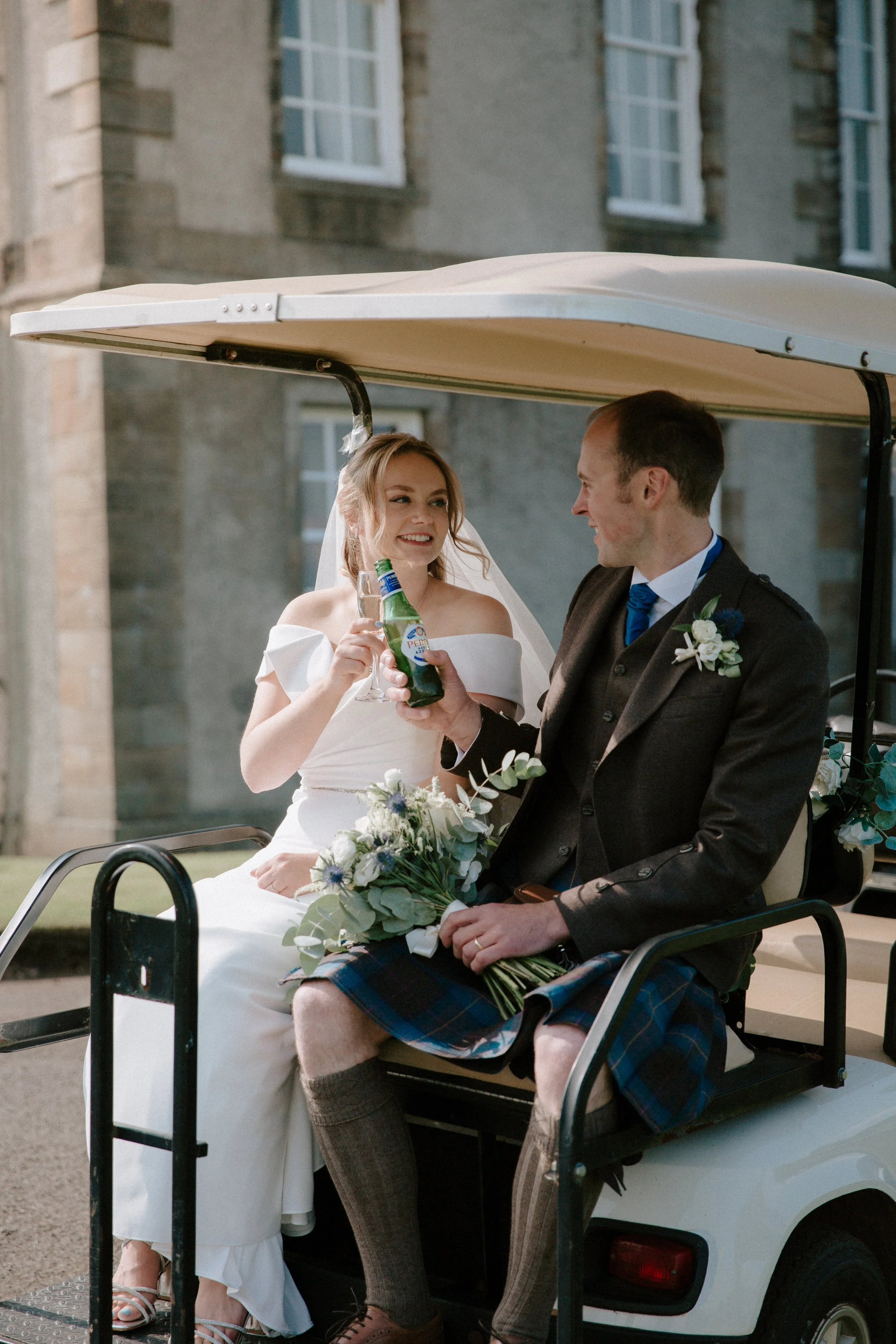 A bride and groom sitting on a golf cart, celebrating with drinks. The bride is holding a glass and smiling, while the groom holds a beer bottle, both dressed in wedding attire with a church or historic building in the background.