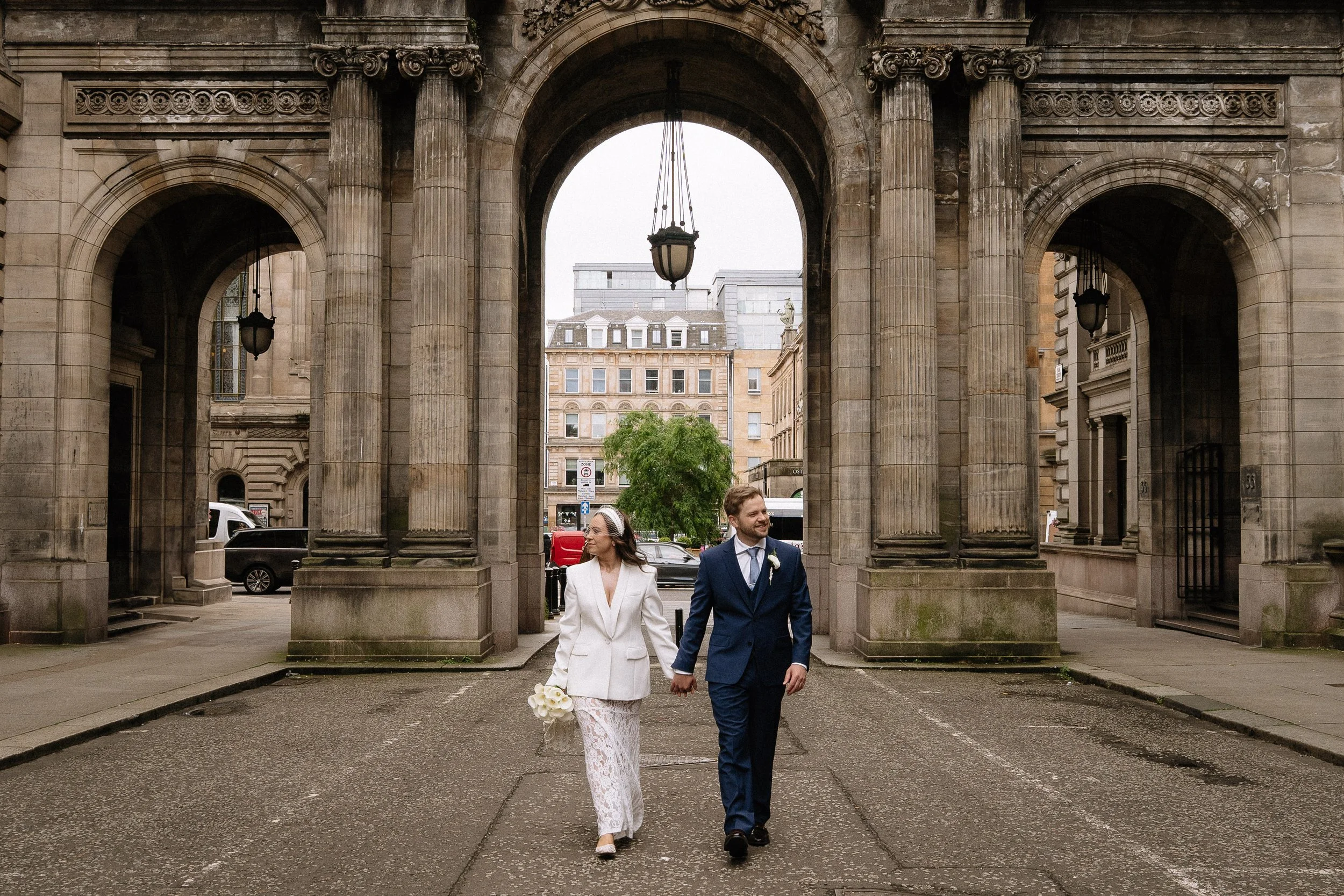 A newlywed couple walking hand-in-hand through an urban archway. The bride is holding white roses, wearing a white lace wedding dress with a white blazer, and a veil. The groom is dressed in a navy suit with a white shirt and blue tie. The setting fe
