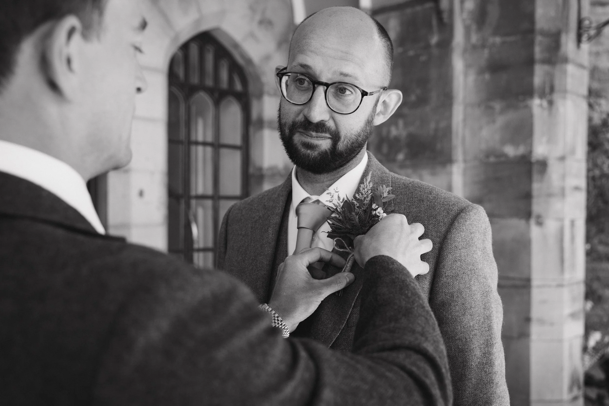 A man with glasses and a beard is pinning a boutonniere onto another man's suit jacket during a wedding or formal event, with a stone building and arched windows in the background. - captured by an Edinburgh wedding photographer