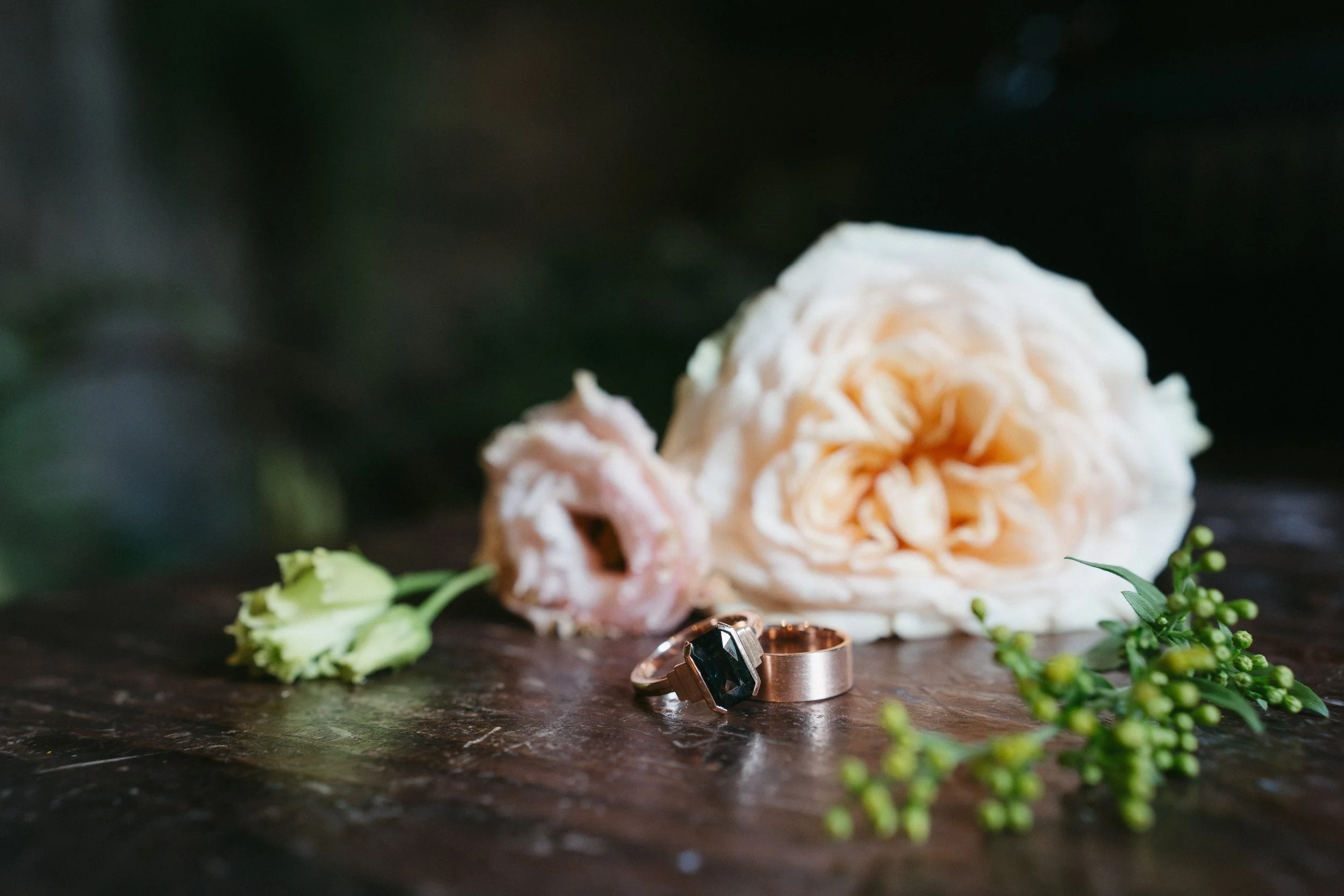 A collection of rings, flowers, and a large white rose on a wooden surface with a blurred dark background - captured by an Edinburgh wedding photographer