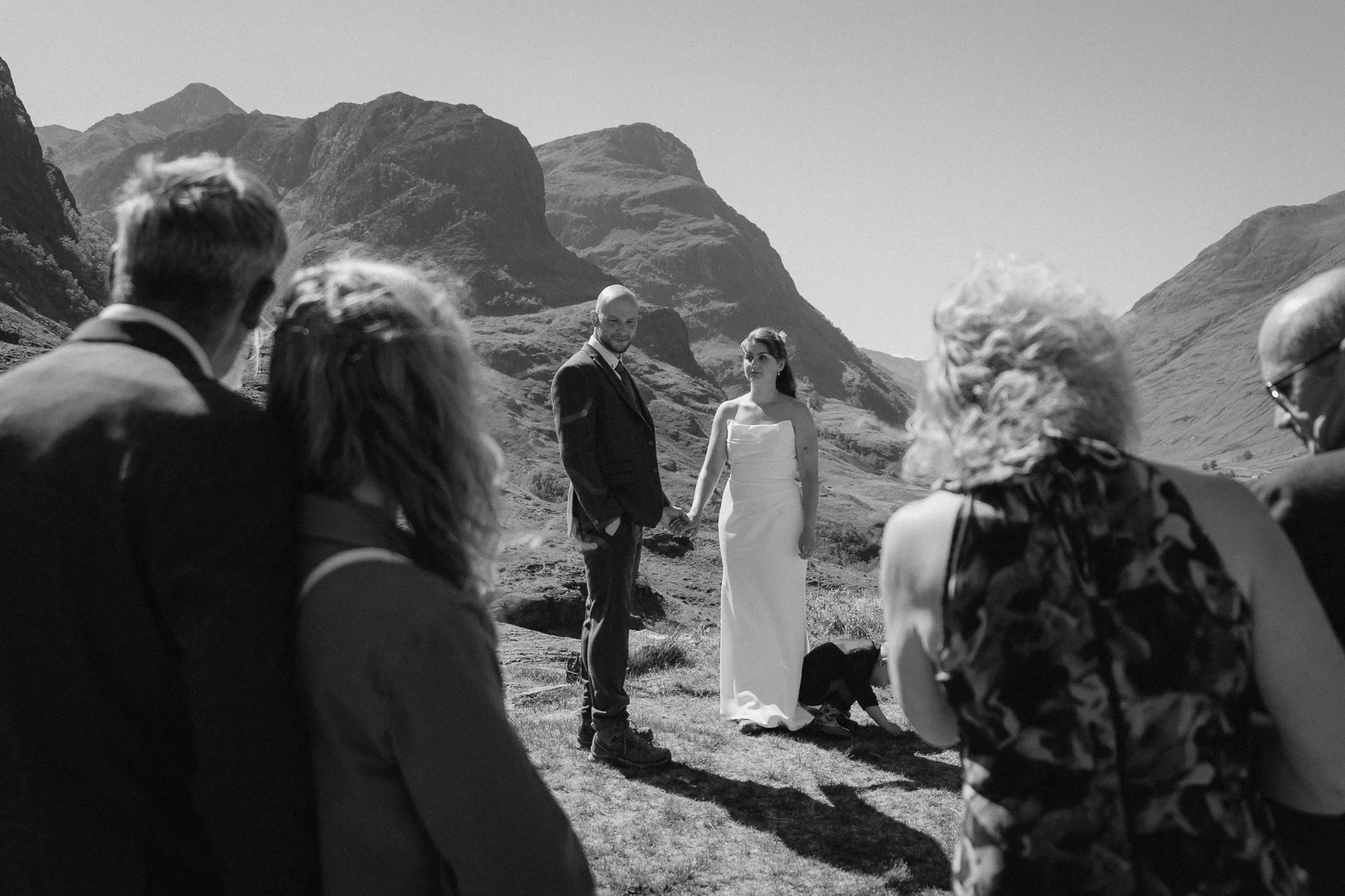 A black and white photo of a wedding ceremony outdoors with the couple holding hands and standing in front of a mountain landscape, surrounded by guests. - captured by an Edinburgh wedding photographer