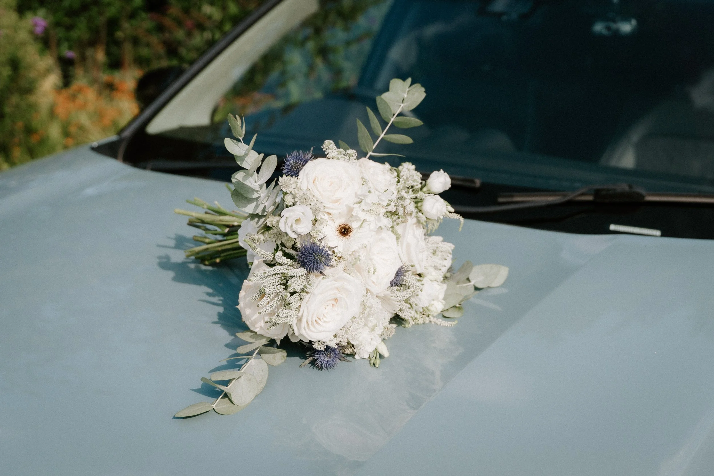 A bouquet of white flowers with various greenery is placed on the hood of a light-colored car. - captured by an Edinburgh wedding photographer