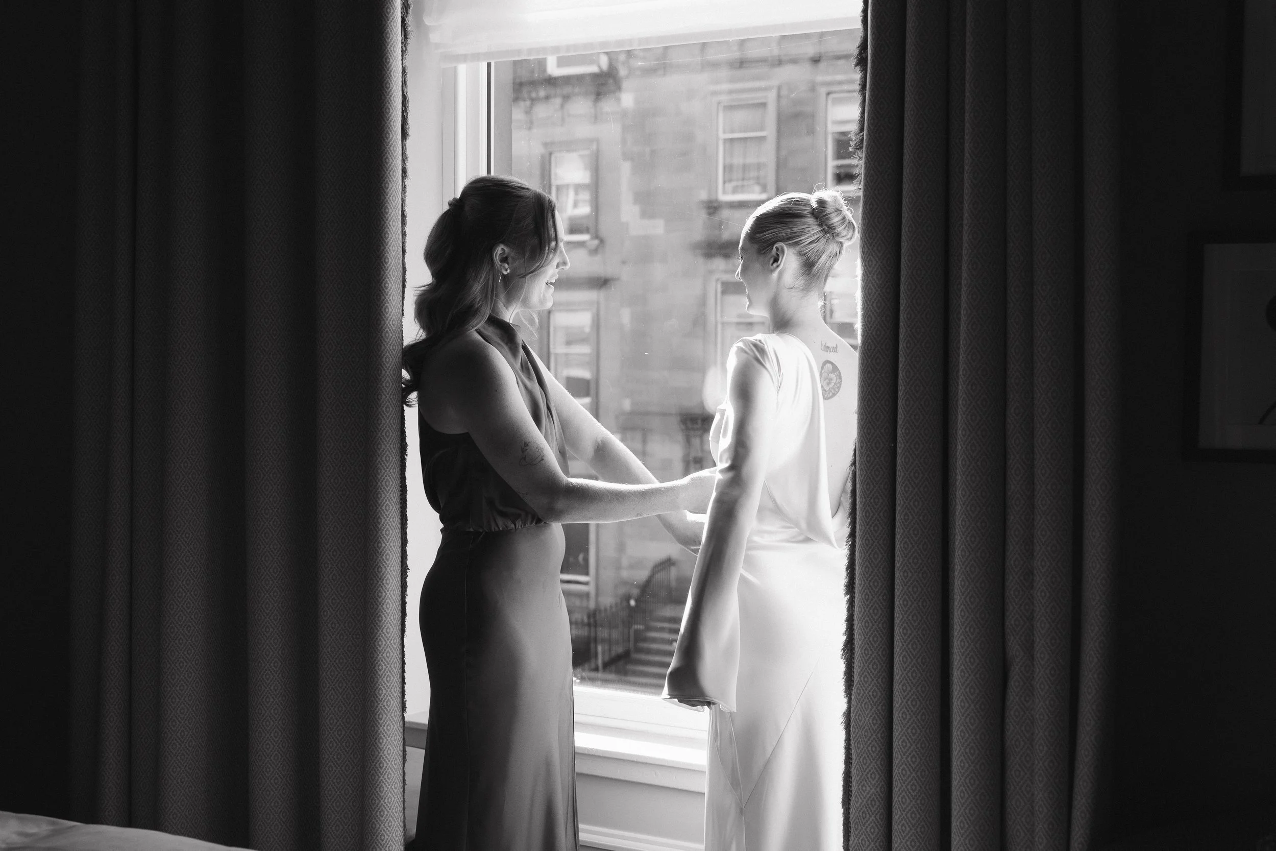 Two women holding hands and looking at each other by a large window, with curtains on either side, in black and white. - captured by an Edinburgh wedding photographer