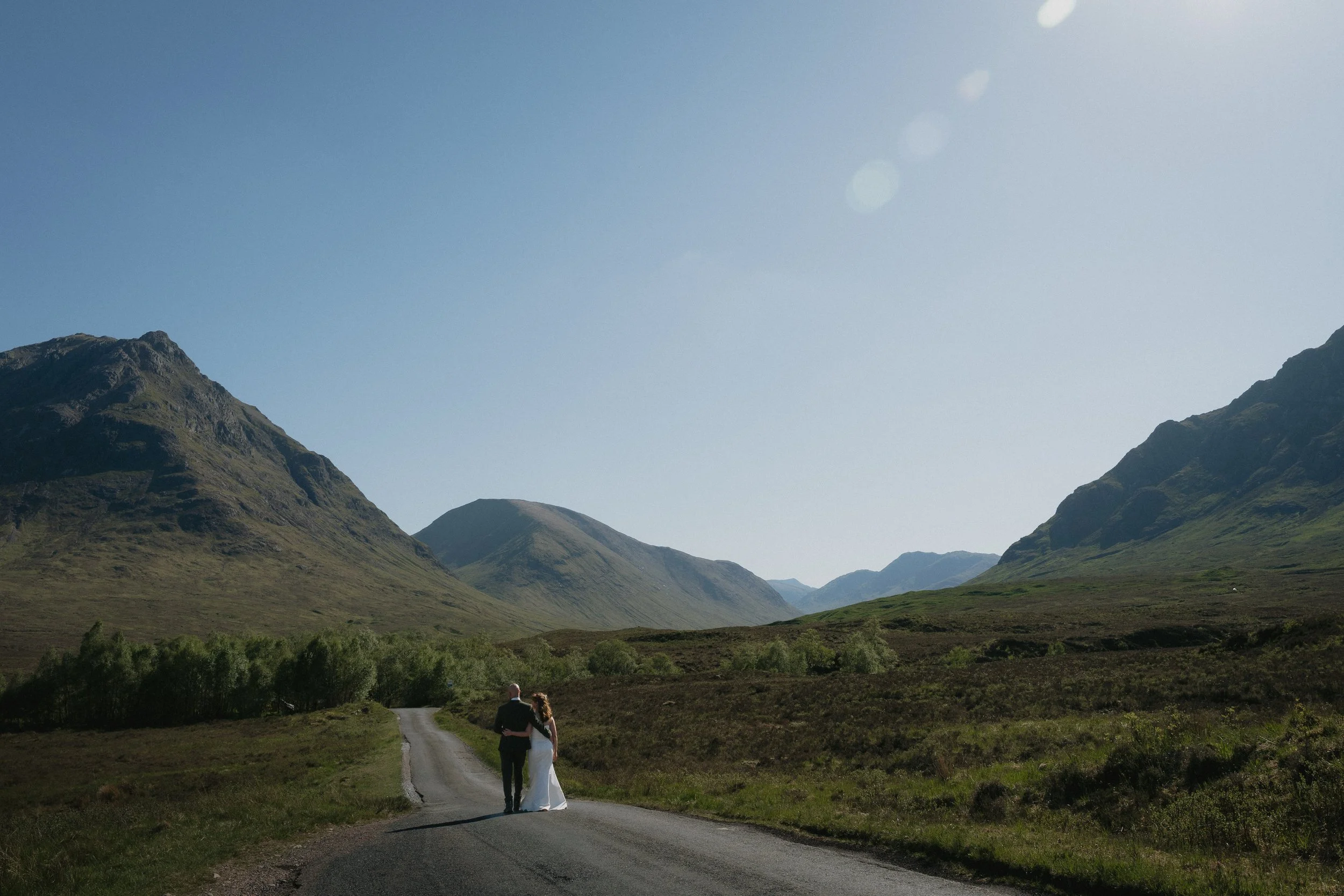 A couple in wedding attire walking along a deserted country road in a mountainous landscape under a clear blue sky. - captured by an Edinburgh wedding photographer