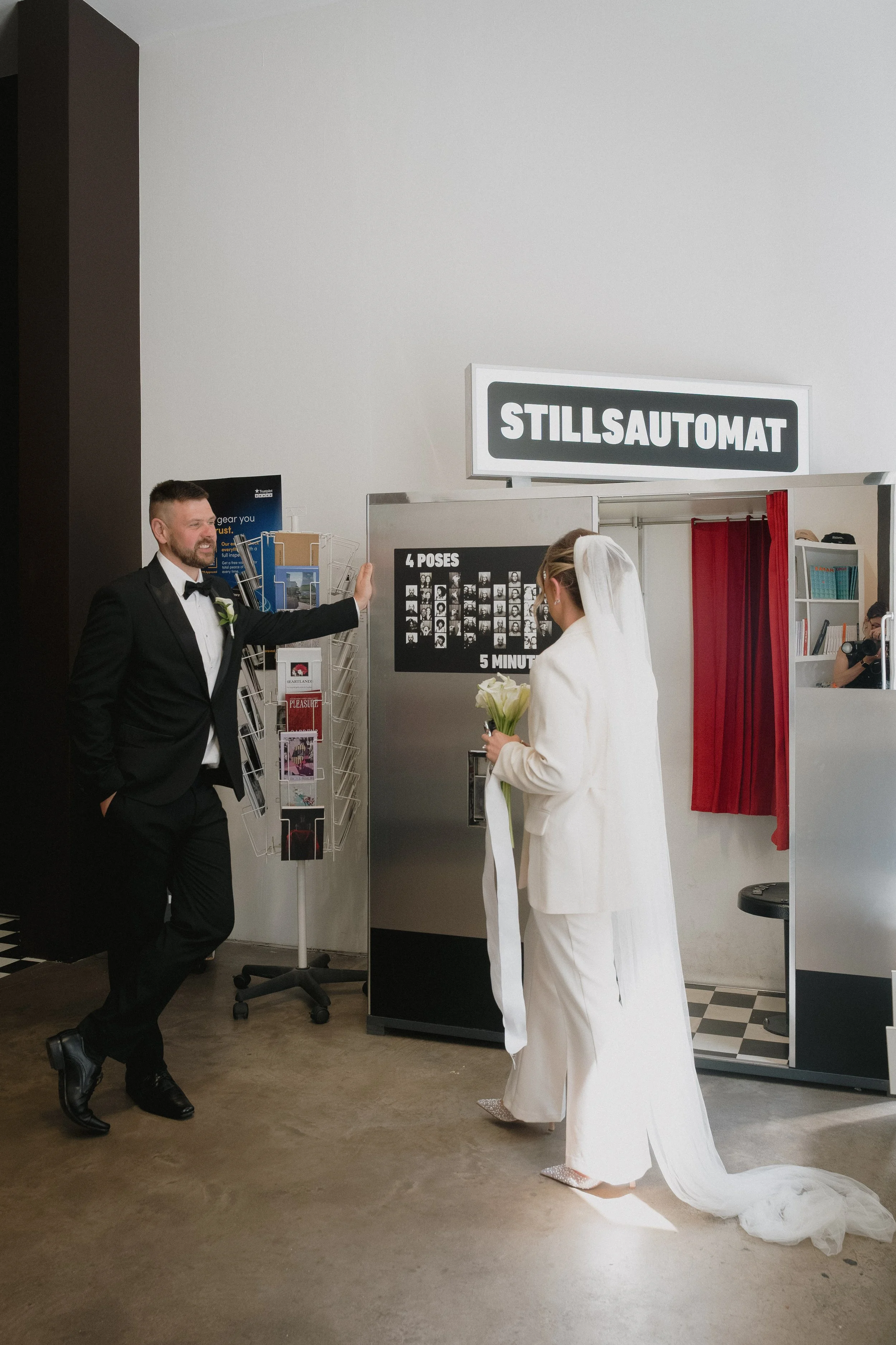 A bride in a white wedding dress and veil holding a bouquet of white roses, standing in front of a photo booth with a sign labeled 'STILLSAUTOMAT'. A groom in a black tuxedo is talking to her, leaning against a rack of postcards or tourist brochures.