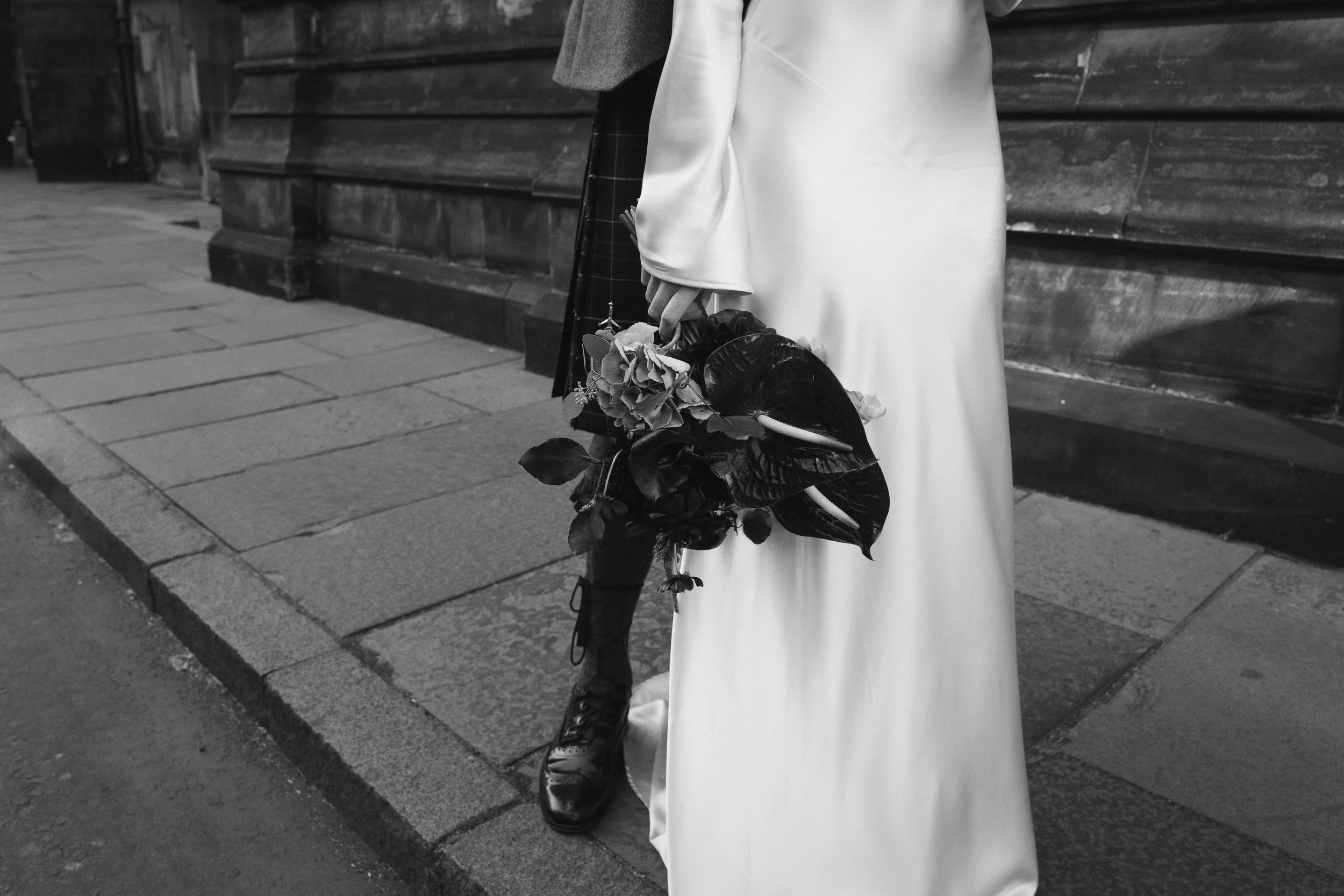 Person in a long white dress holding a bouquet of flowers and wearing black boots, standing on a sidewalk next to a wooden wall. - captured by an Edinburgh wedding photographer