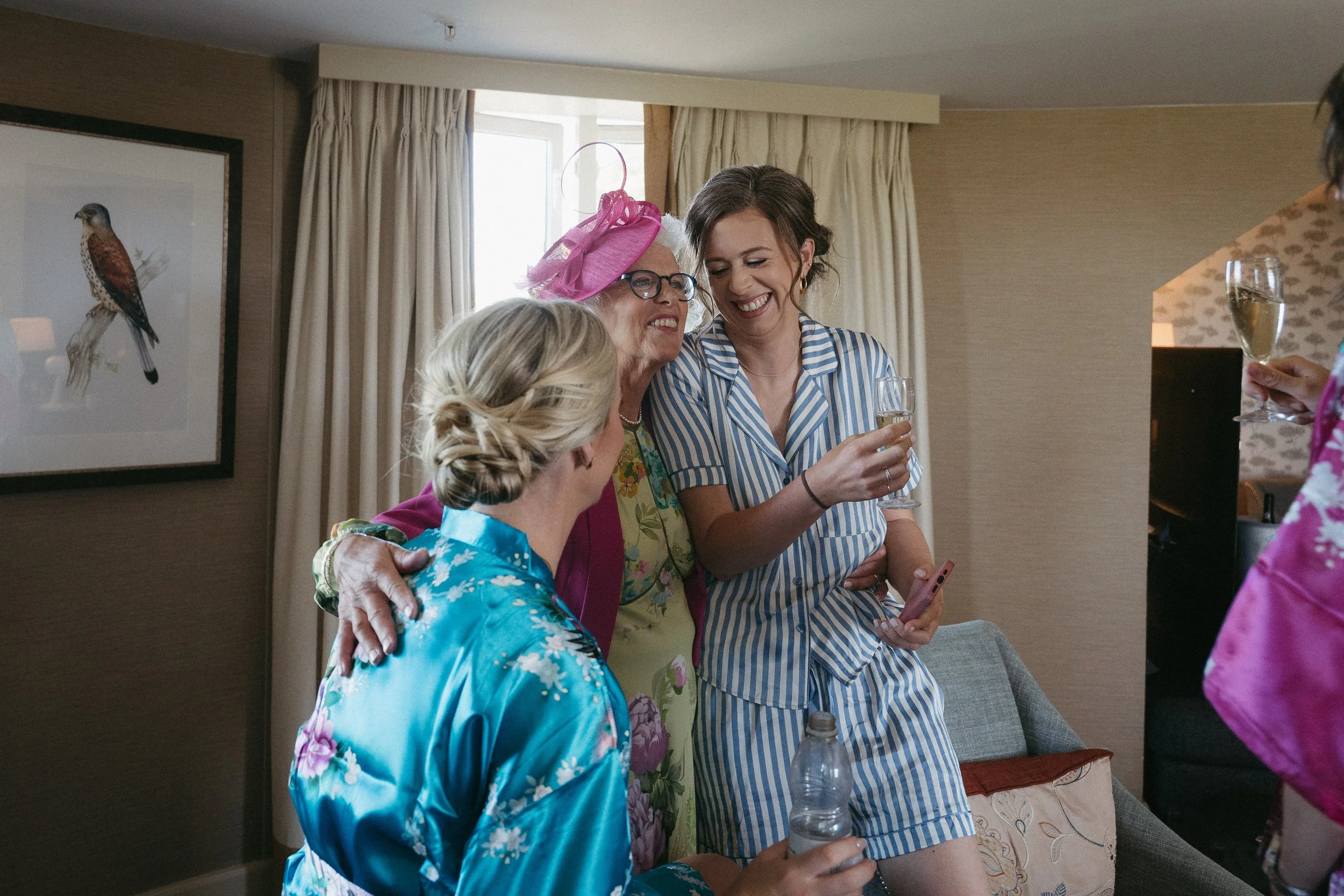 Four women in colorful pajamas celebrating together indoors, with one woman wearing a pink fascinator hat and another holding a bottle of water, all smiling and holding glasses of champagne. - captured by an Edinburgh wedding photographer