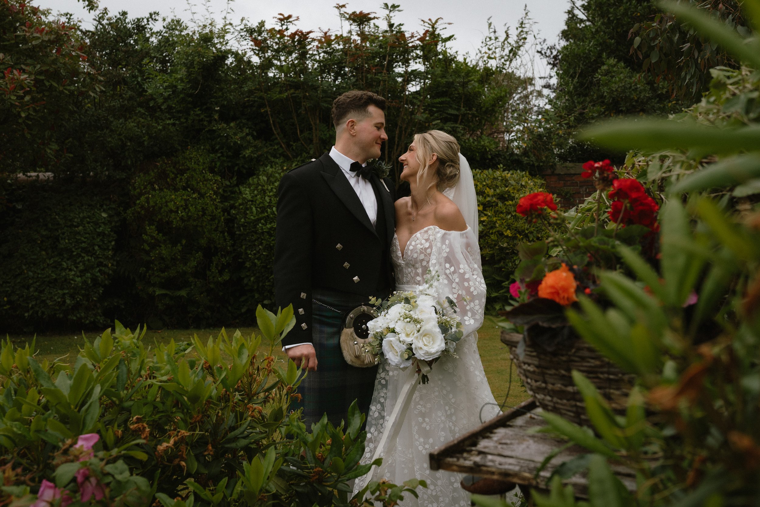 A bride and groom in wedding attire standing close together in a garden, smiling and looking into each other's eyes, with lush greenery and flowers around them. - captured by an Edinburgh wedding photographer