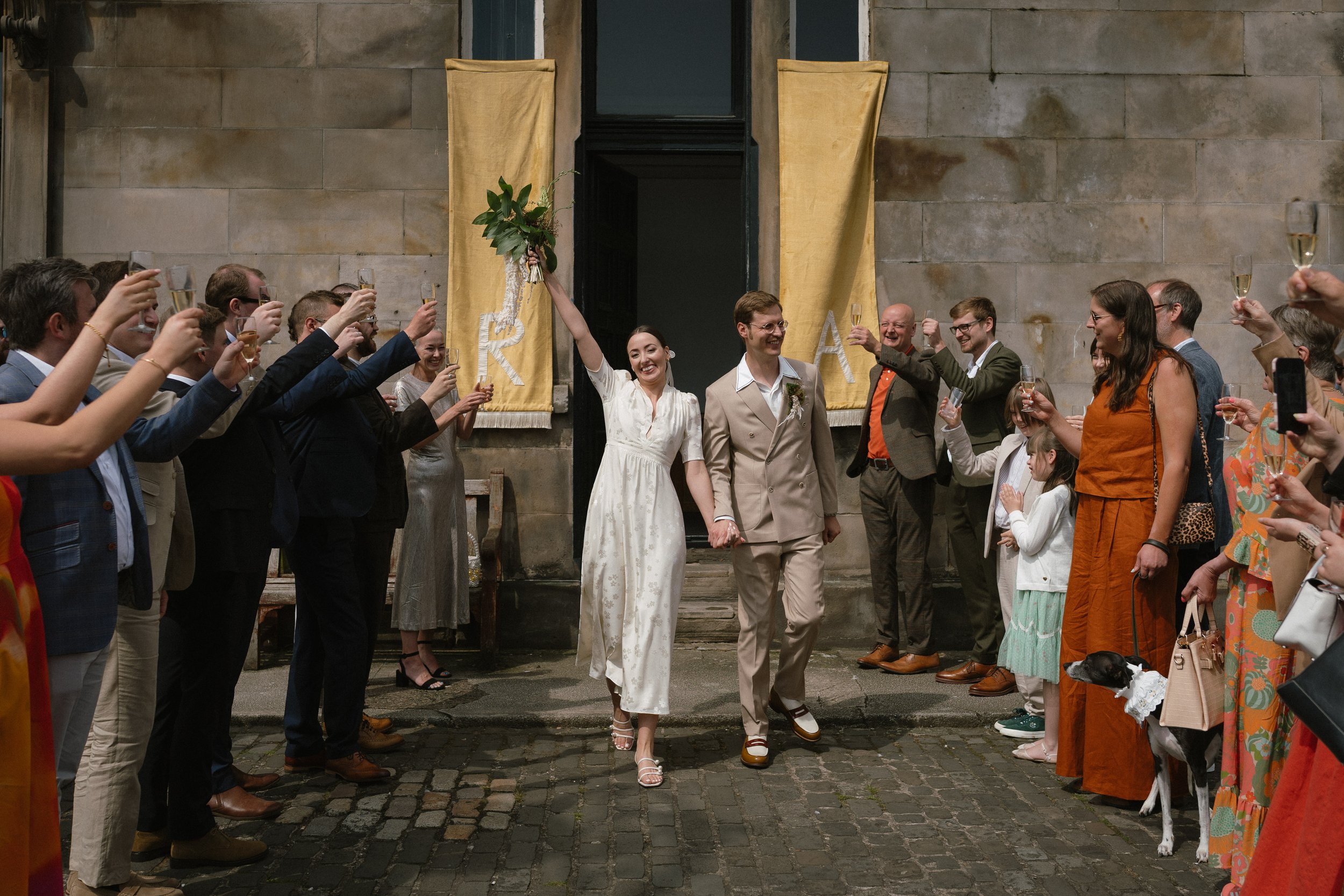 A newlywed couple walking hand in hand while guests cheer and toast with glasses of champagne outside a building with stone walls and yellow banners. - captured by an Edinburgh wedding photographer