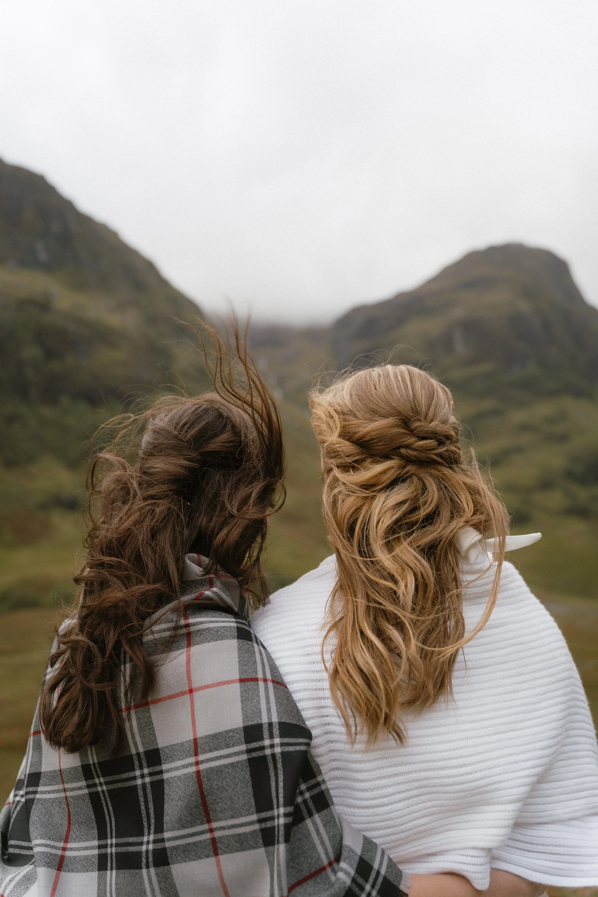 Two women with tousled hair, one with blonde and the other with brown hair, standing outdoors on a misty day, with green hills and mountains in the background - captured by an Edinburgh wedding photographer