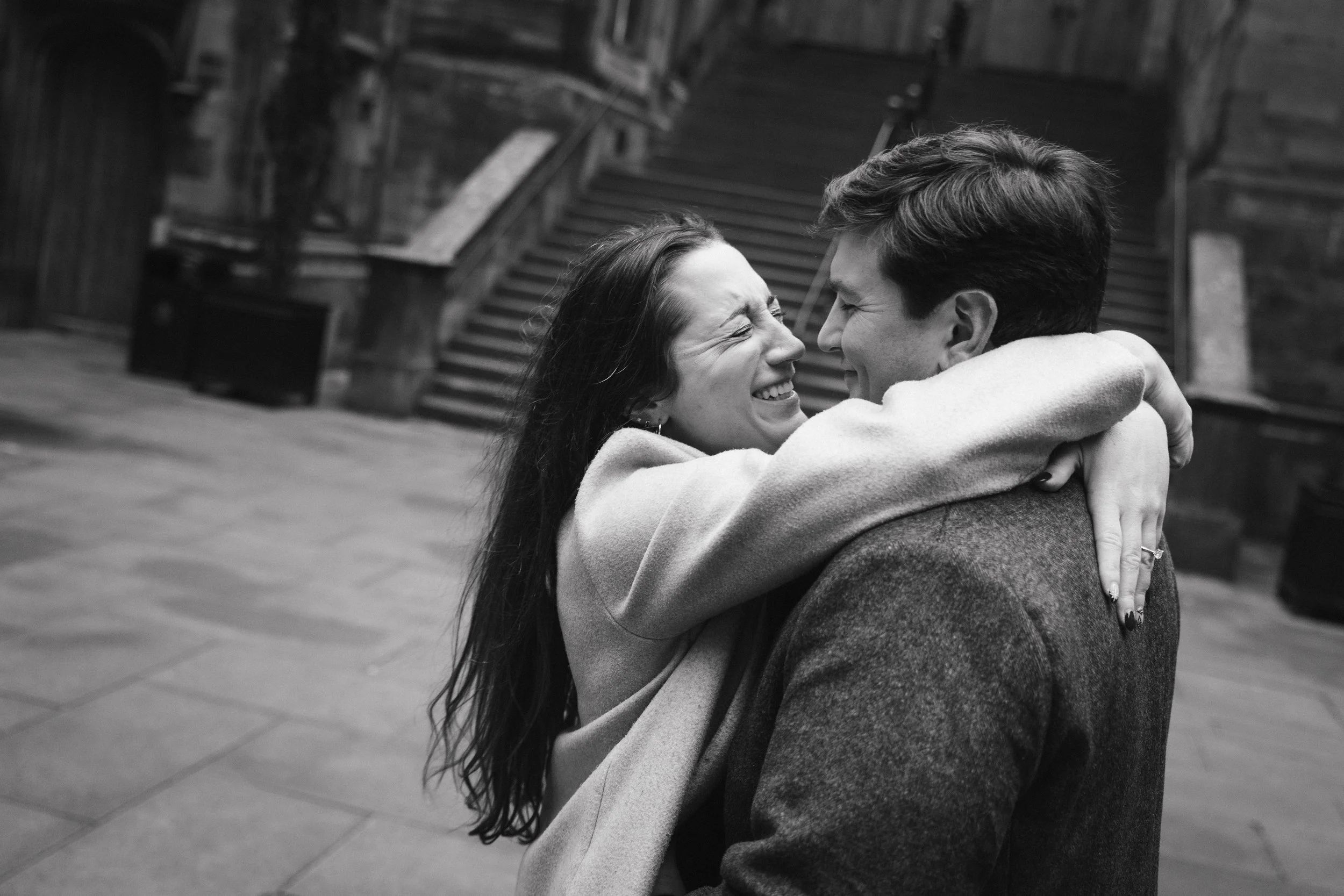 A couple embracing and smiling joyfully in a black-and-white photo, with stairs in the background.
