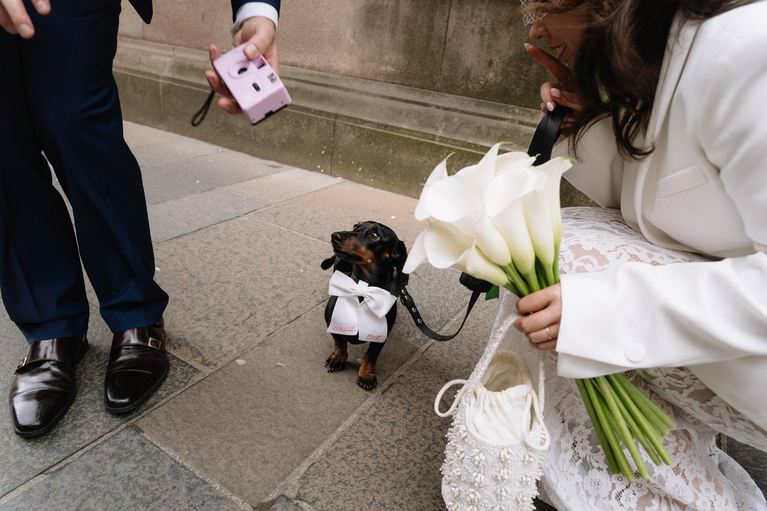 A woman in a white jacket holding a bouquet of white calla lilies crouches down near a small black dog wearing a white bow tie and a white coat, while a person nearby holds a pink instant camera.