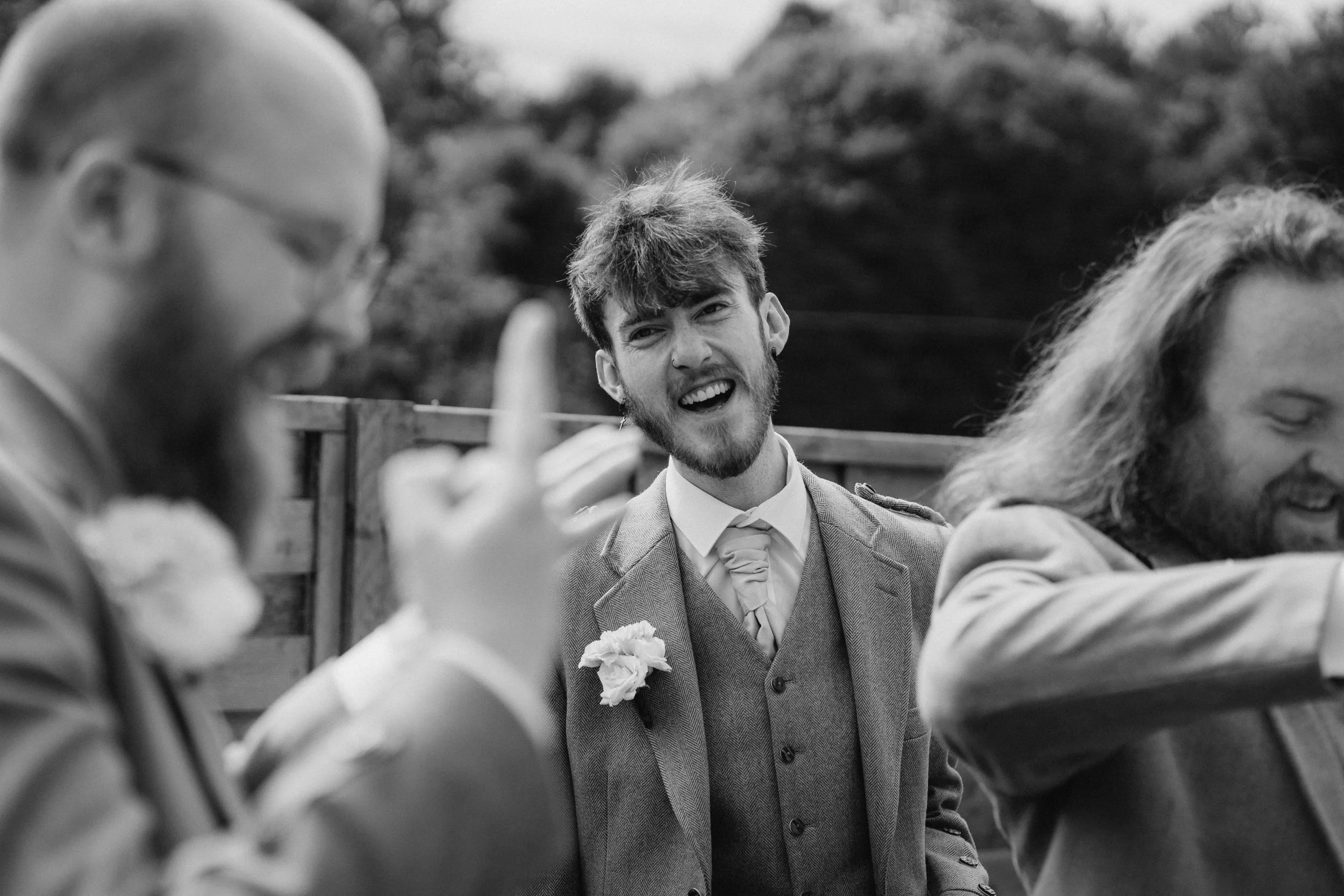 A black and white photograph showing three men at an outdoor event. The man in the center is smiling and wearing a suit with a boutonniere, suggesting a wedding or formal gathering. The other two men, one on each side, are blurred and appear to be en