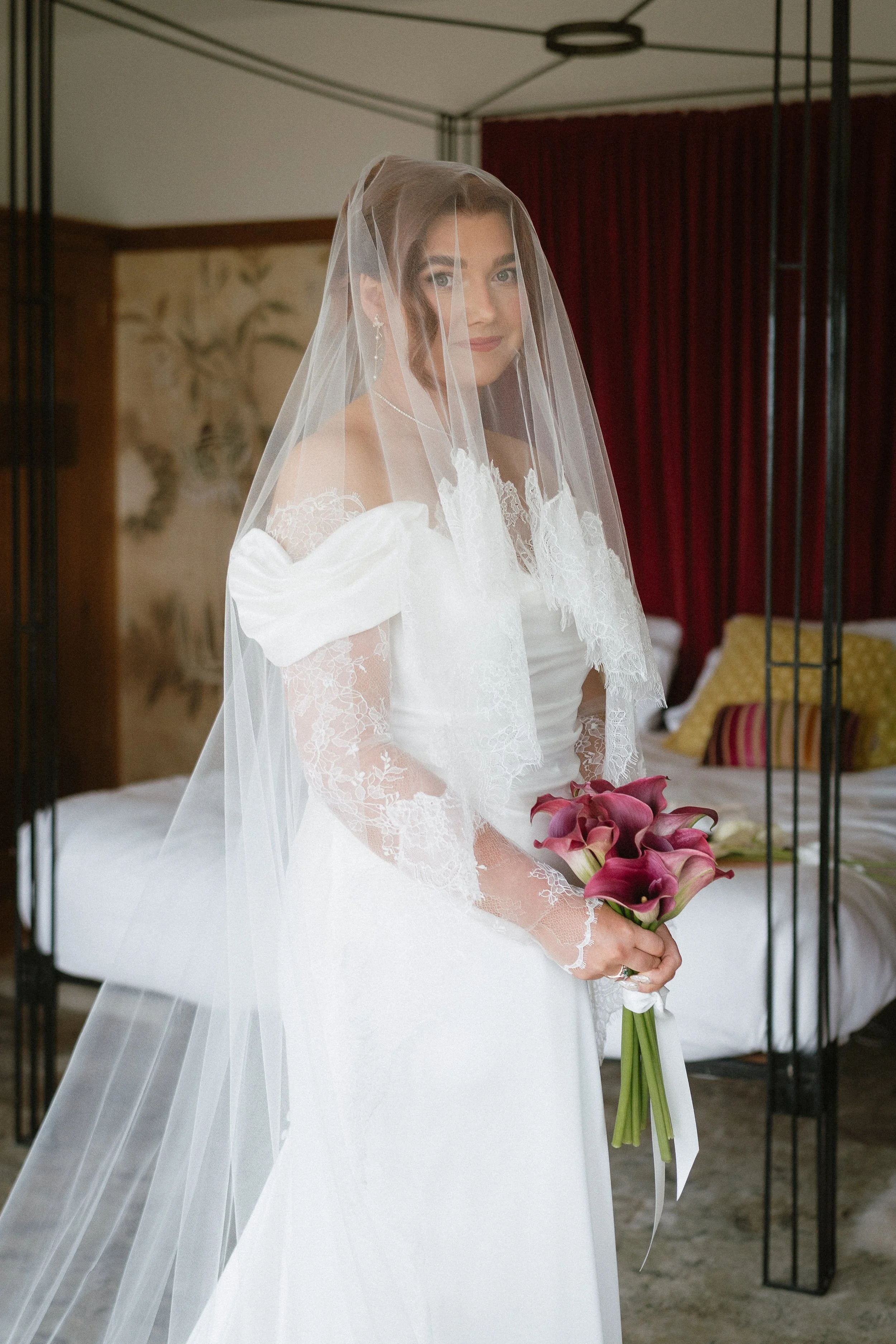 A bride in a white wedding dress with lace sleeves and a long veil holding a bouquet of pink calla lilies, standing in a room with a bed and red curtains. - captured by an Edinburgh wedding photographer