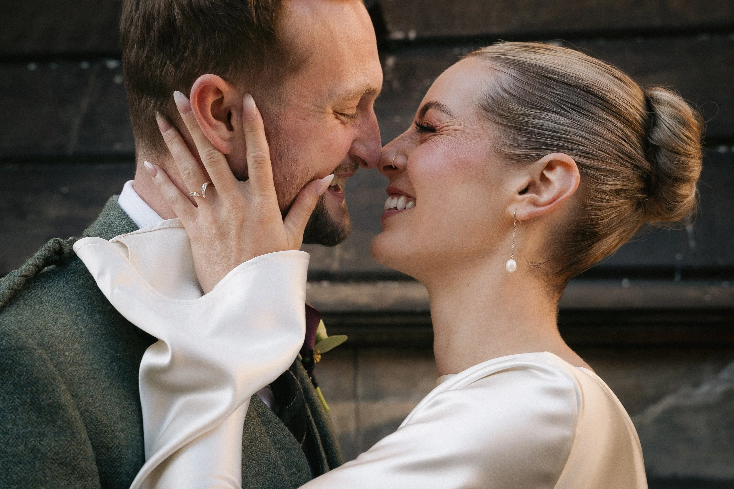 A couple in wedding attire touching noses and smiling, with the woman holding the man's face. The woman has a pearl earring and a ring; the man has a beard and short hair.