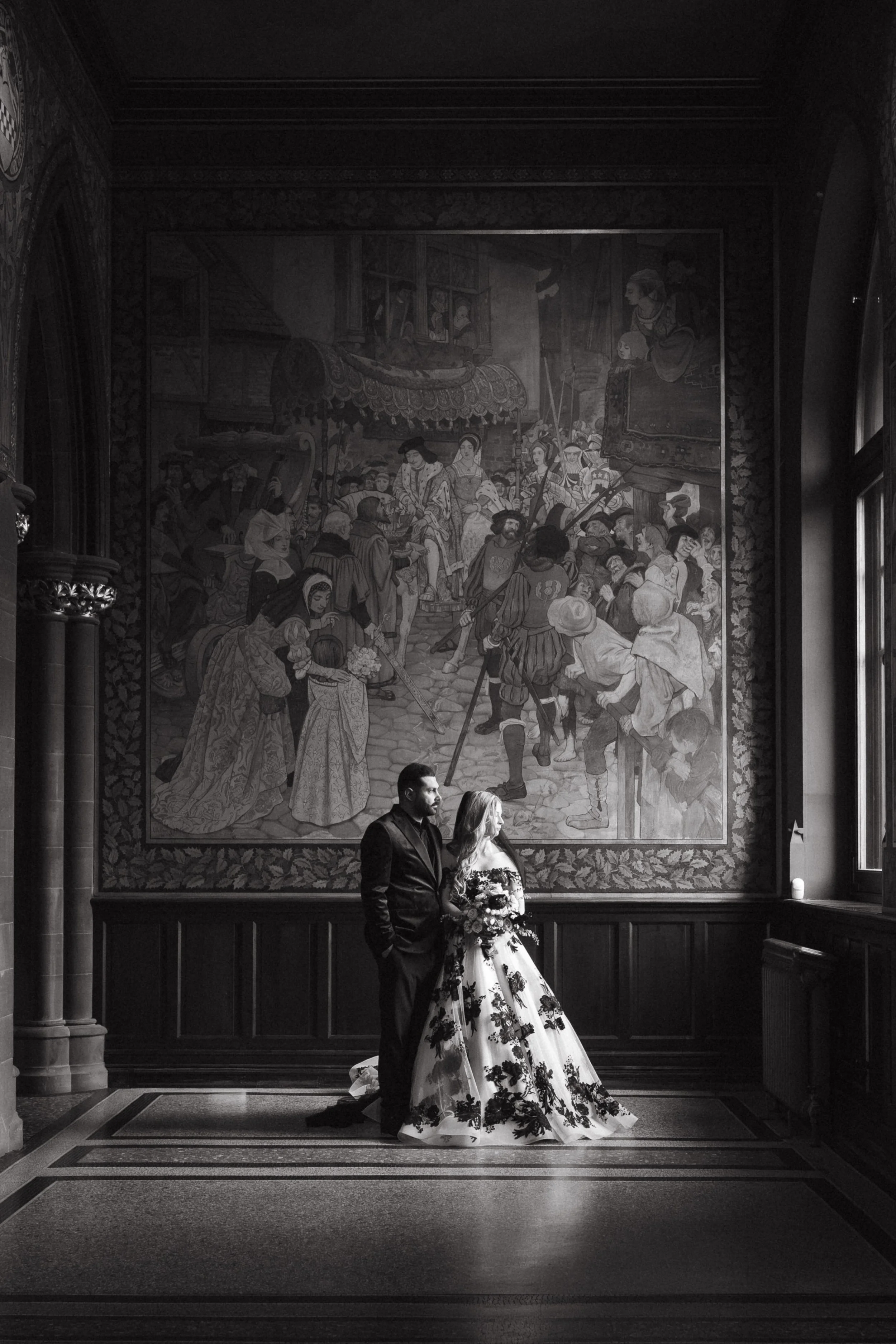 A couple in wedding attire standing inside a grand hall, with a large historical painting on the wall behind them, illuminated by natural light from nearby windows. - captured by an Edinburgh wedding photographer