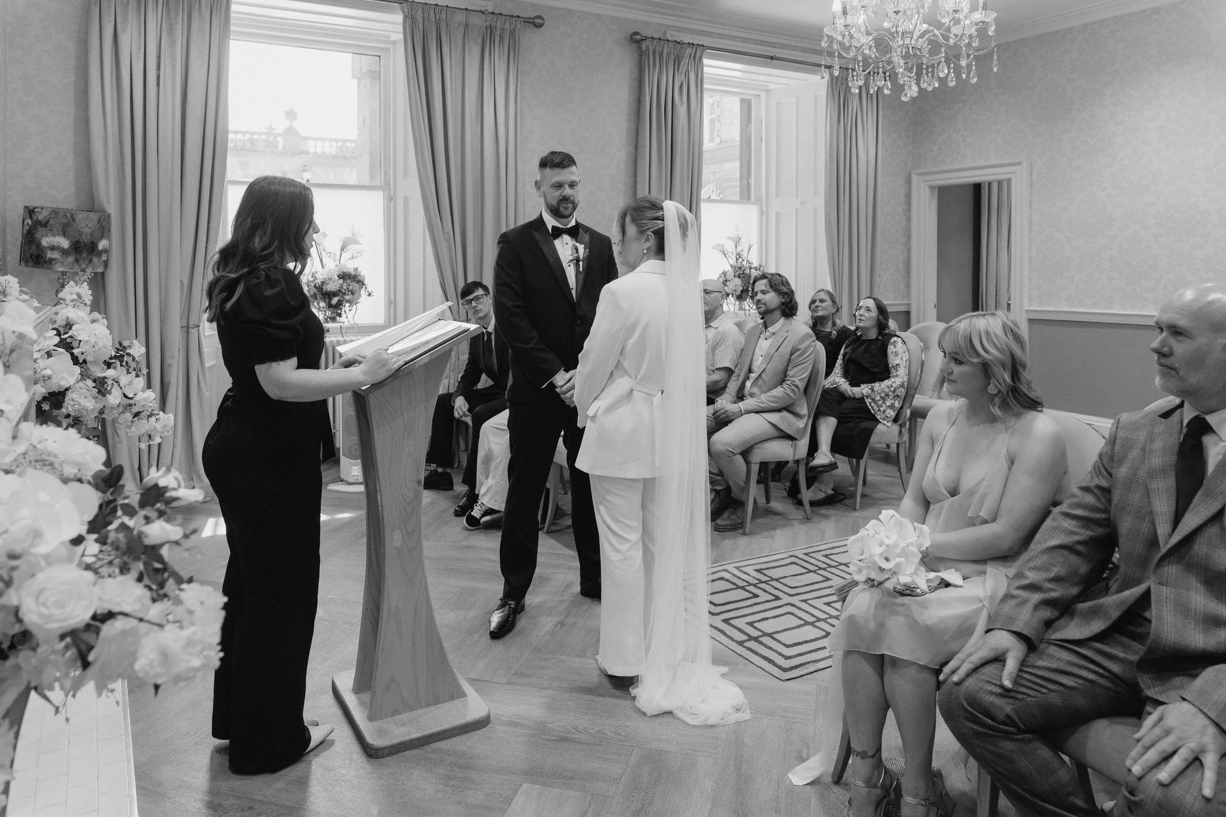 A black and white photo of a wedding ceremony with a bride and groom facing each other and holding hands, an officiant reading from a book, guests seated in the background, and floral arrangements visible.