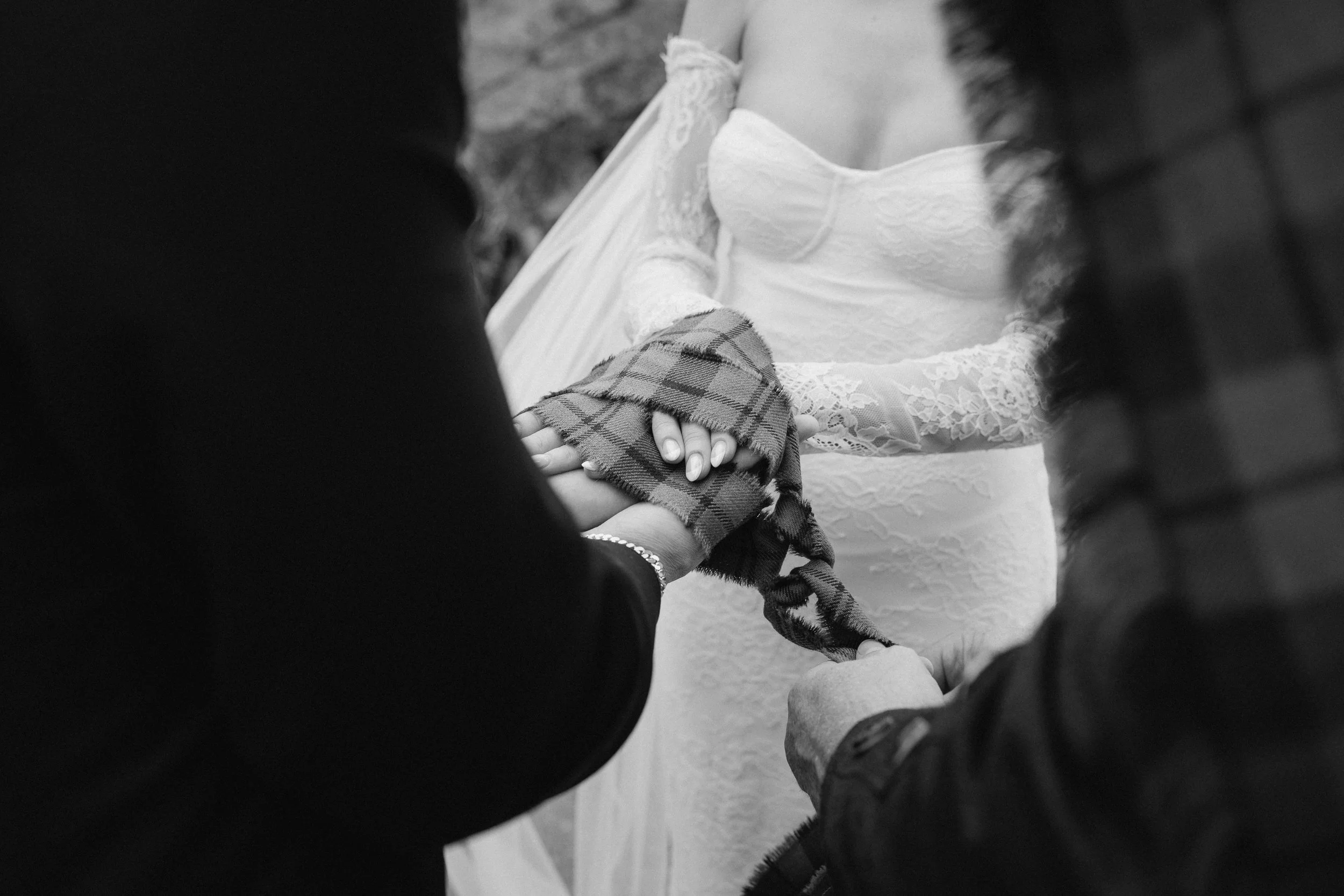 A black and white photo of a couple holding hands during a wedding ceremony, with the bride in a lace wedding dress and the groom in a dark suit, stylishly holding her hand wrapped in a plaid cloth. - captured by an Edinburgh wedding photographer