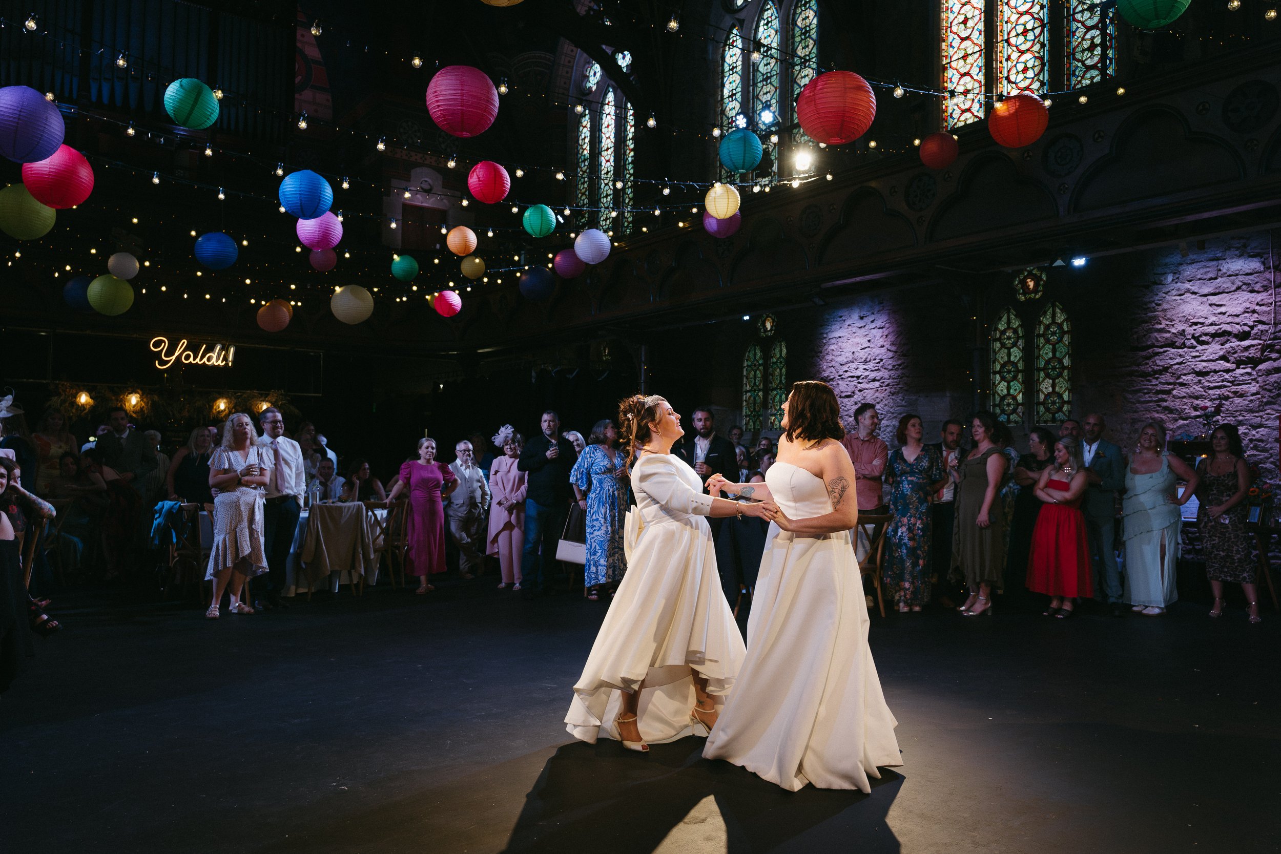 Two brides dancing at their wedding reception in a decorated indoor venue with colorful lanterns and string lights. - captured by an Edinburgh wedding photographer